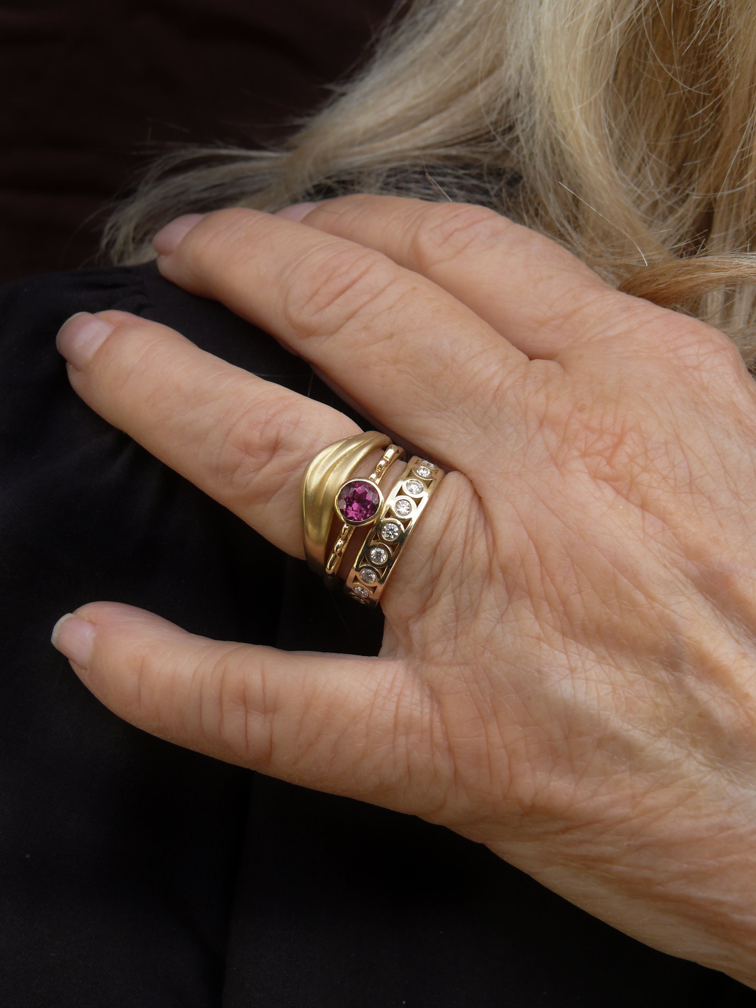 hand wearing gold curved dune ring and cylinder ring with eternity diamonds and gold vessel ring with garnet on dark background