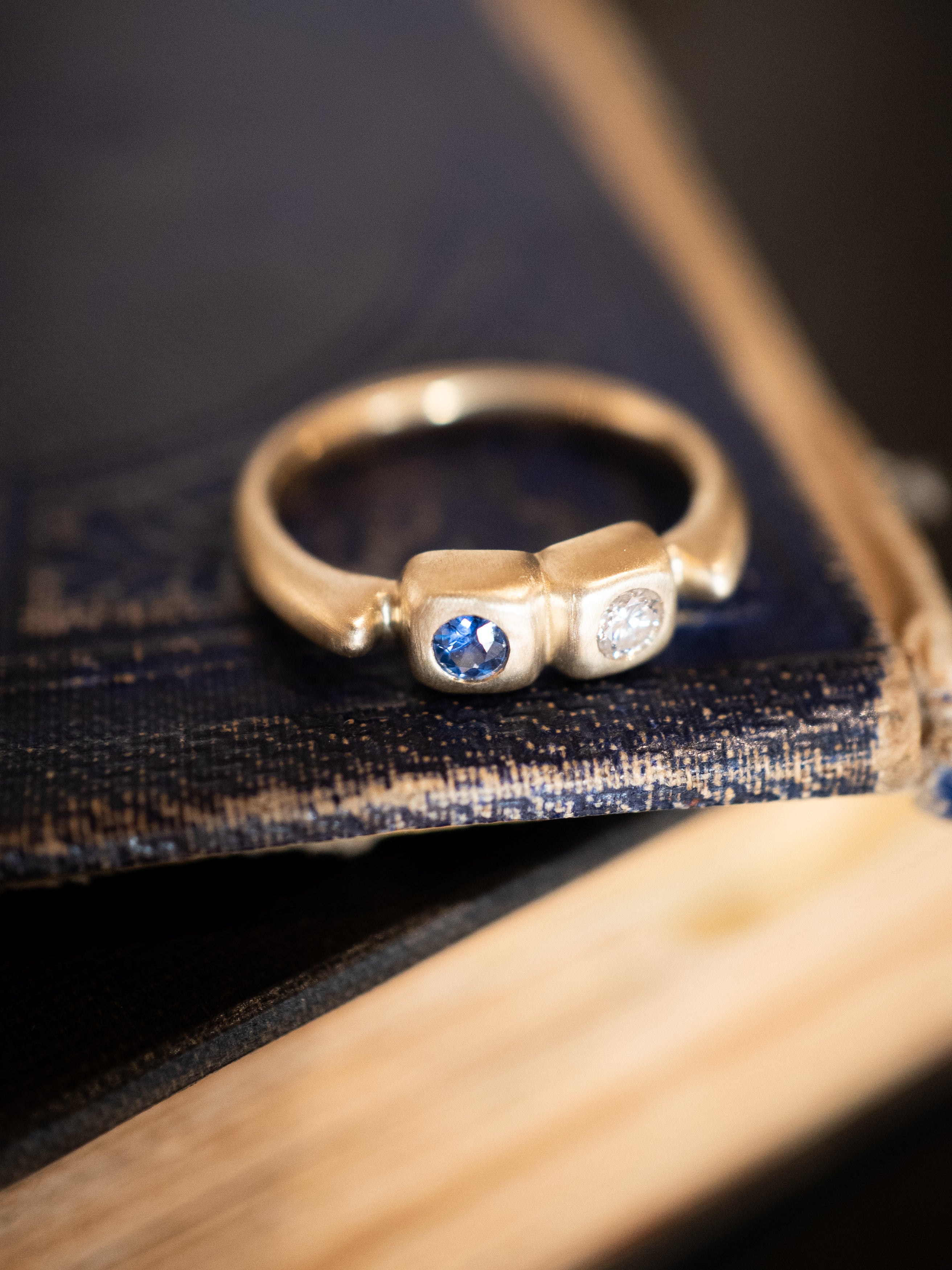 Two-stone gold ring with a blue sapphire and a white diamond on a dark blue background.