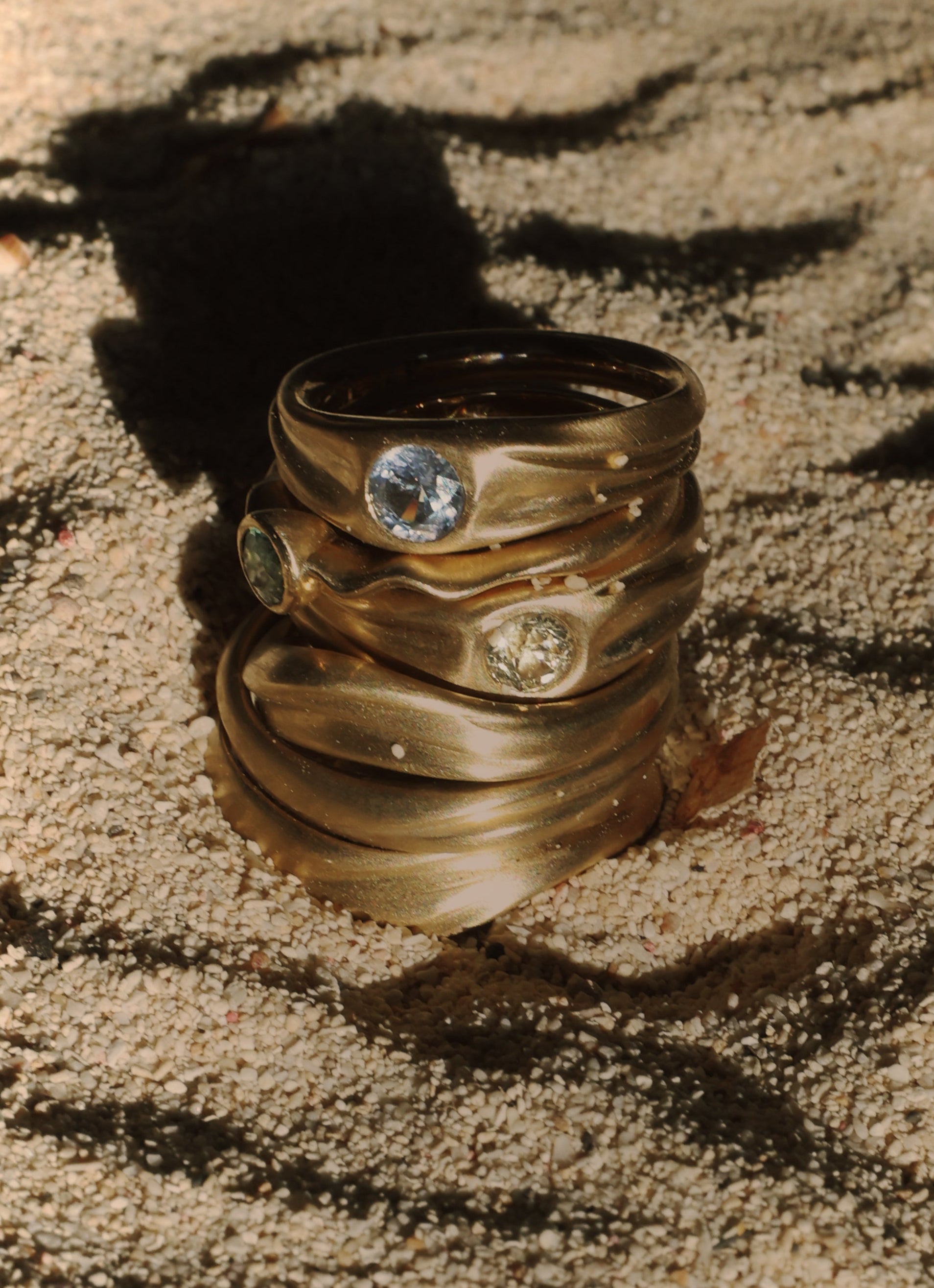 Stack of gold Dune rings with gemstones on a sandy surface