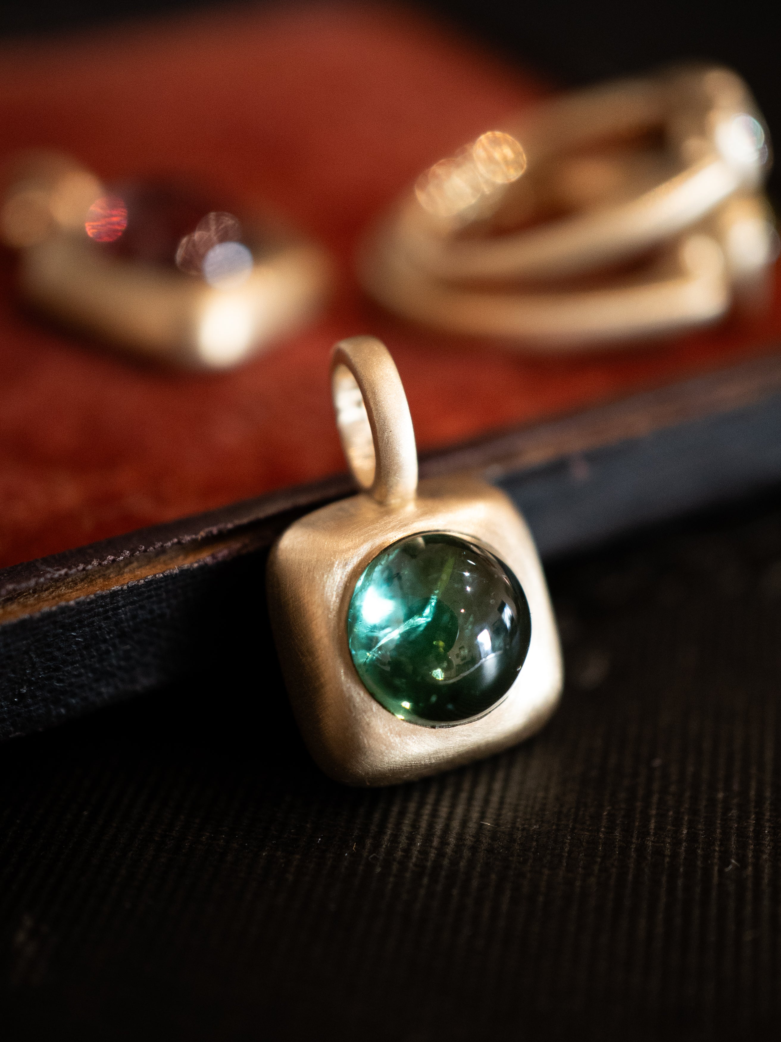 Square gold trove charm with a green tourmaline on a dark surface