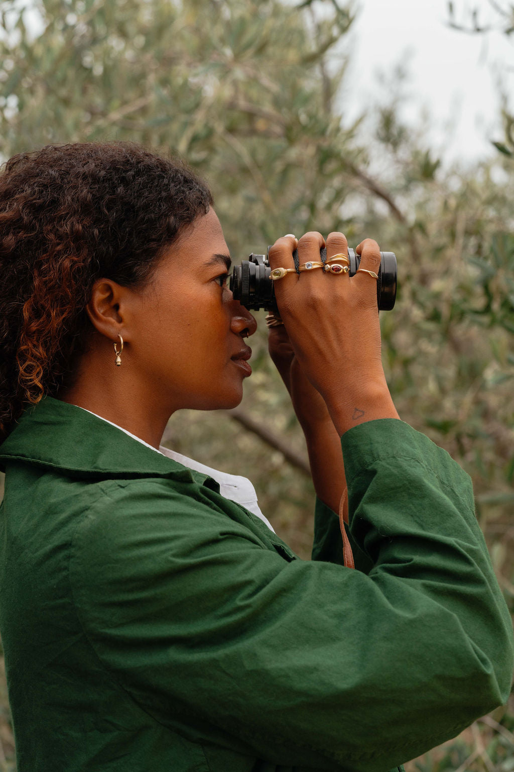 Woman wearing green jacket in olive orchard and looking through binoculars and wearing trove collection gold and gemstone rings and earrings