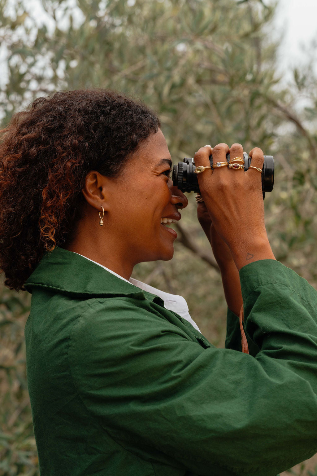 Person in a green jacket using binoculars in an olive orchard wearing gold rings and earrings from Made Line Jewelry Trove Collection.