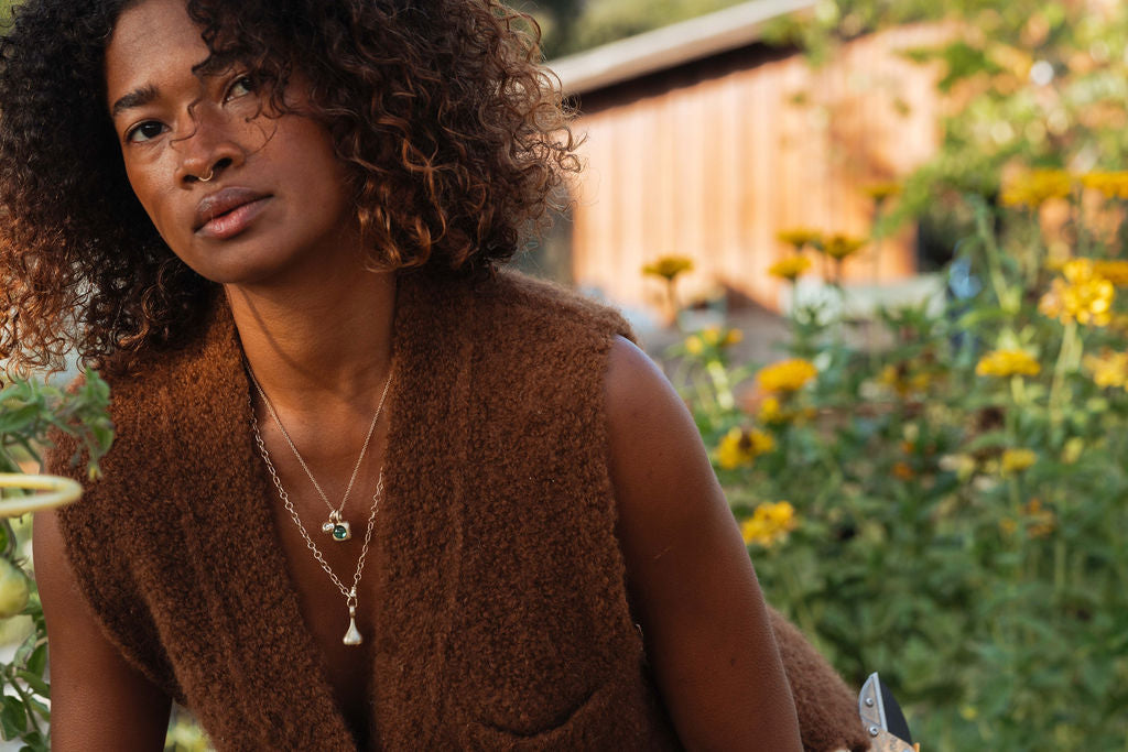 Woman in a brown vest standing in a garden with flowers and a wooden fence in the background, wearing gold chains and charms with bright green tourmaline and citrine pendulum charm.