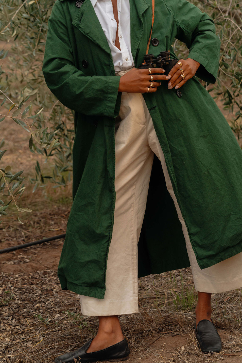 Person wearing a green coat over a white shirt with beige pants, standing outdoors holding onto binoculars and wearing gold and gemstone rings from the Made Line Jewelry Trove Collection.