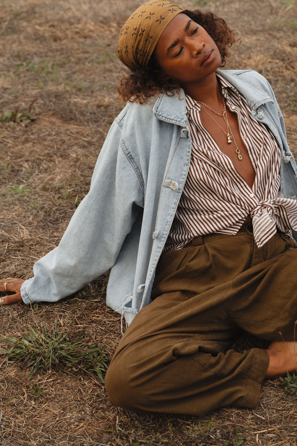 Woman wearing a denim jacket, striped shirt, and brown pants sitting on the ground, wearing gold and gemstone jewelry from the Made Line Jewelry Trove Collection.