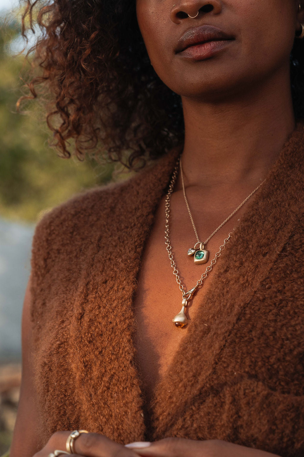 Woman wearing a gold necklace with a green gemstone pendant and a citrine pendant from the Made Line Jewelry Trove Collection, standing outdoors in the garden.