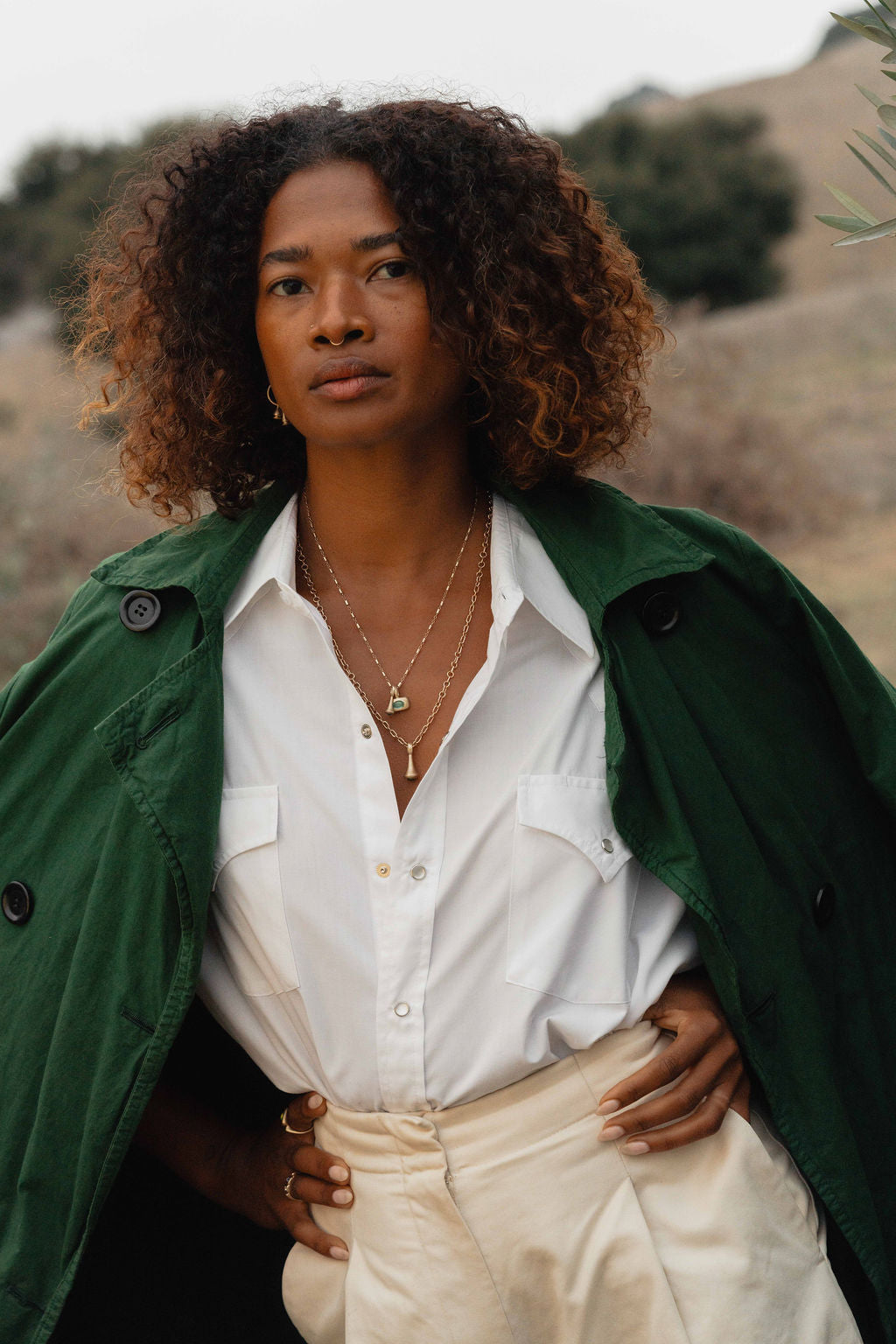 Woman wearing a green jacket over a white shirt with a scenic background, wearing gold pendulum charms and trove charms with green tourmaline