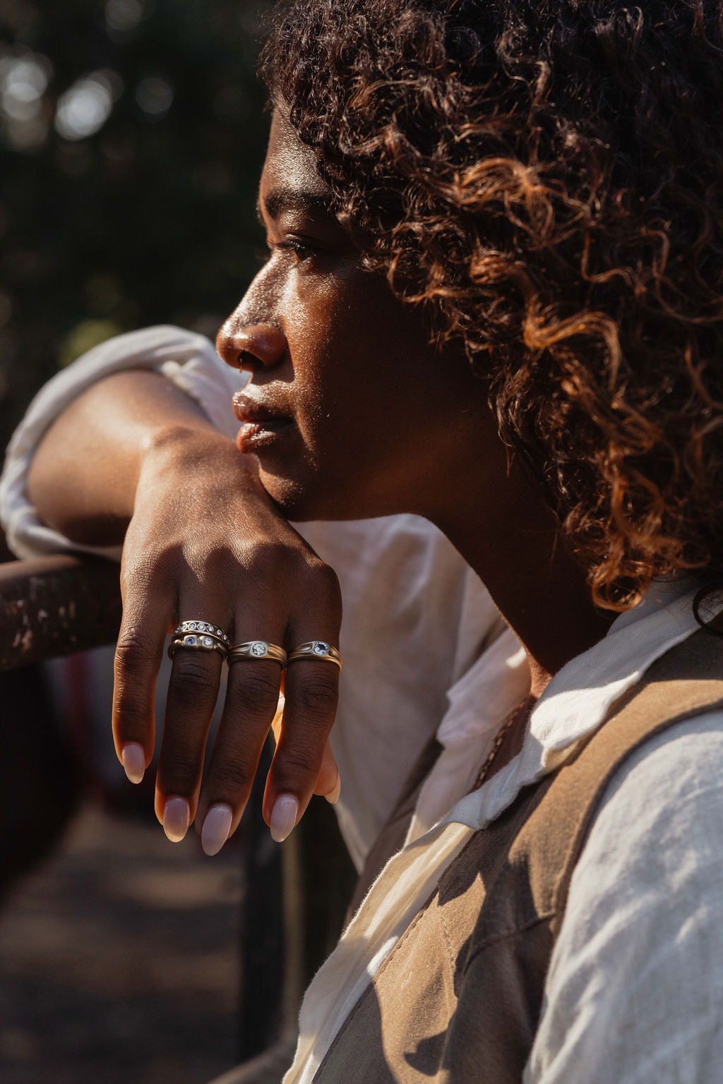 Person with curly hair wearing a white shirt and leather vest with her head resting on her hand with gold rings featuring diamonds, blue sapphires from the Made Line Trove Collection