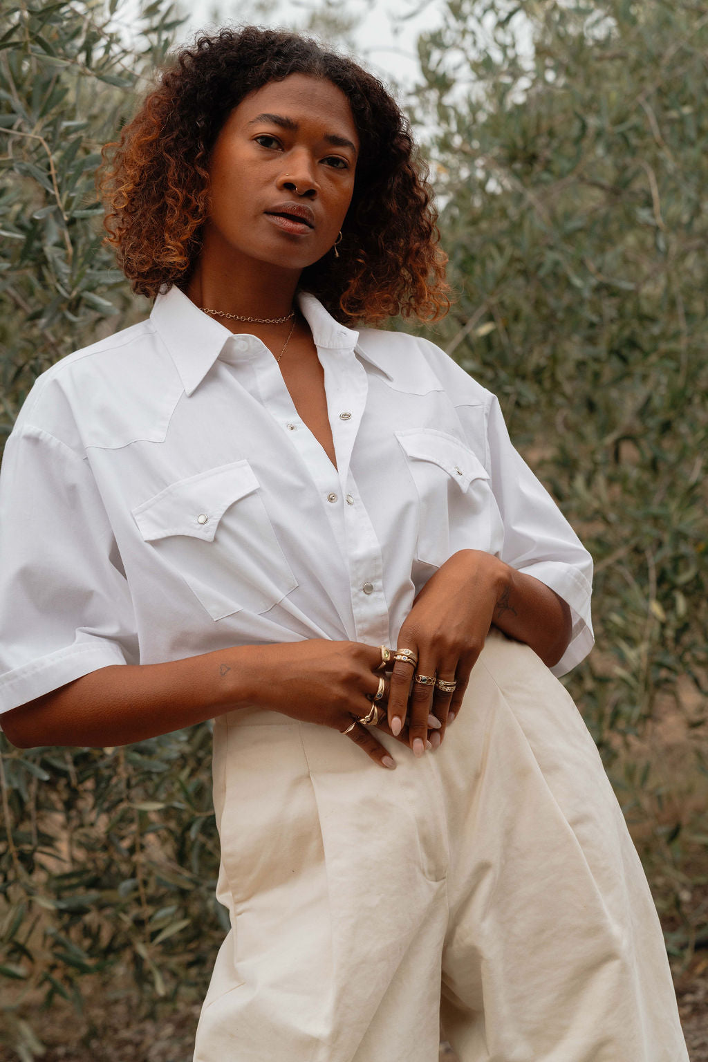 Woman wearing a white shirt and beige pants standing in front of olive trees, wearing gold necklace and rings from Made Line Jewelry Trove Collection.