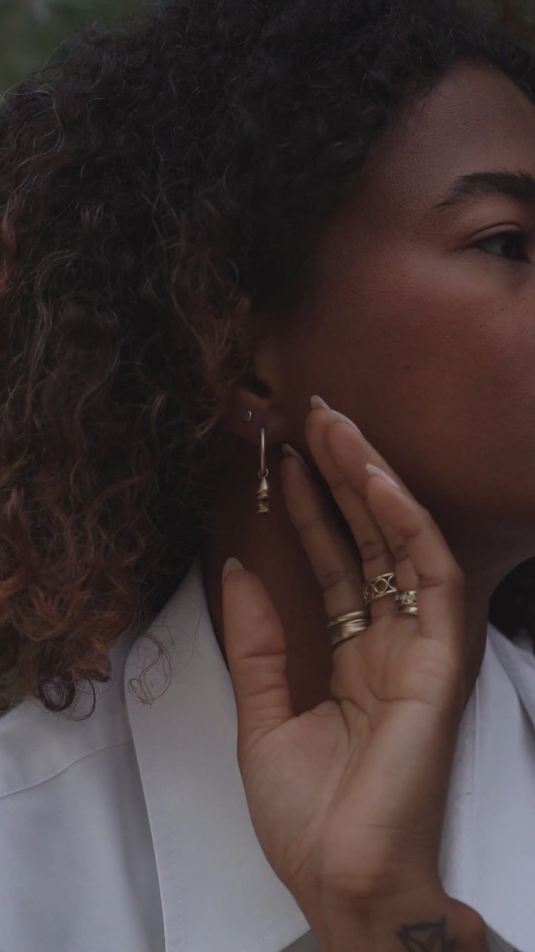 Model with curly hair wearing gold rings and pendulum charm hoop earrings with sapphires and emeralds, from the Made Line Jewelry Trove Collection.