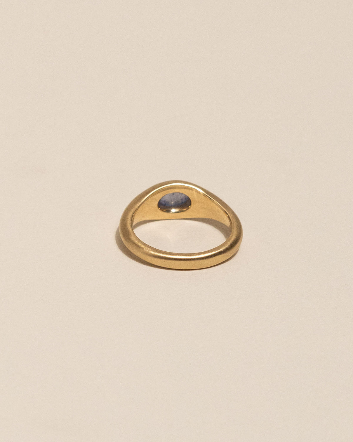 Back view of a gold signet ring with an oval blue stone, centered on a warm beige background.