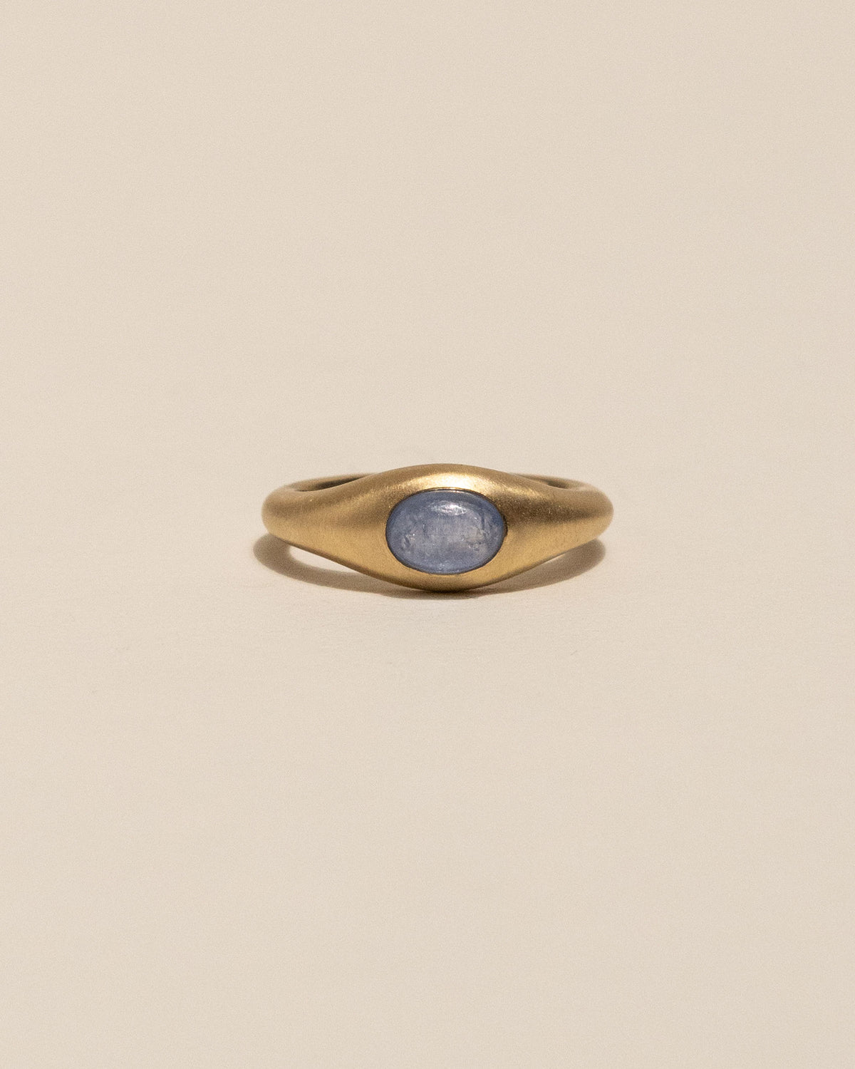 A gold signet ring with an oval blue stone, centered on a warm beige background.