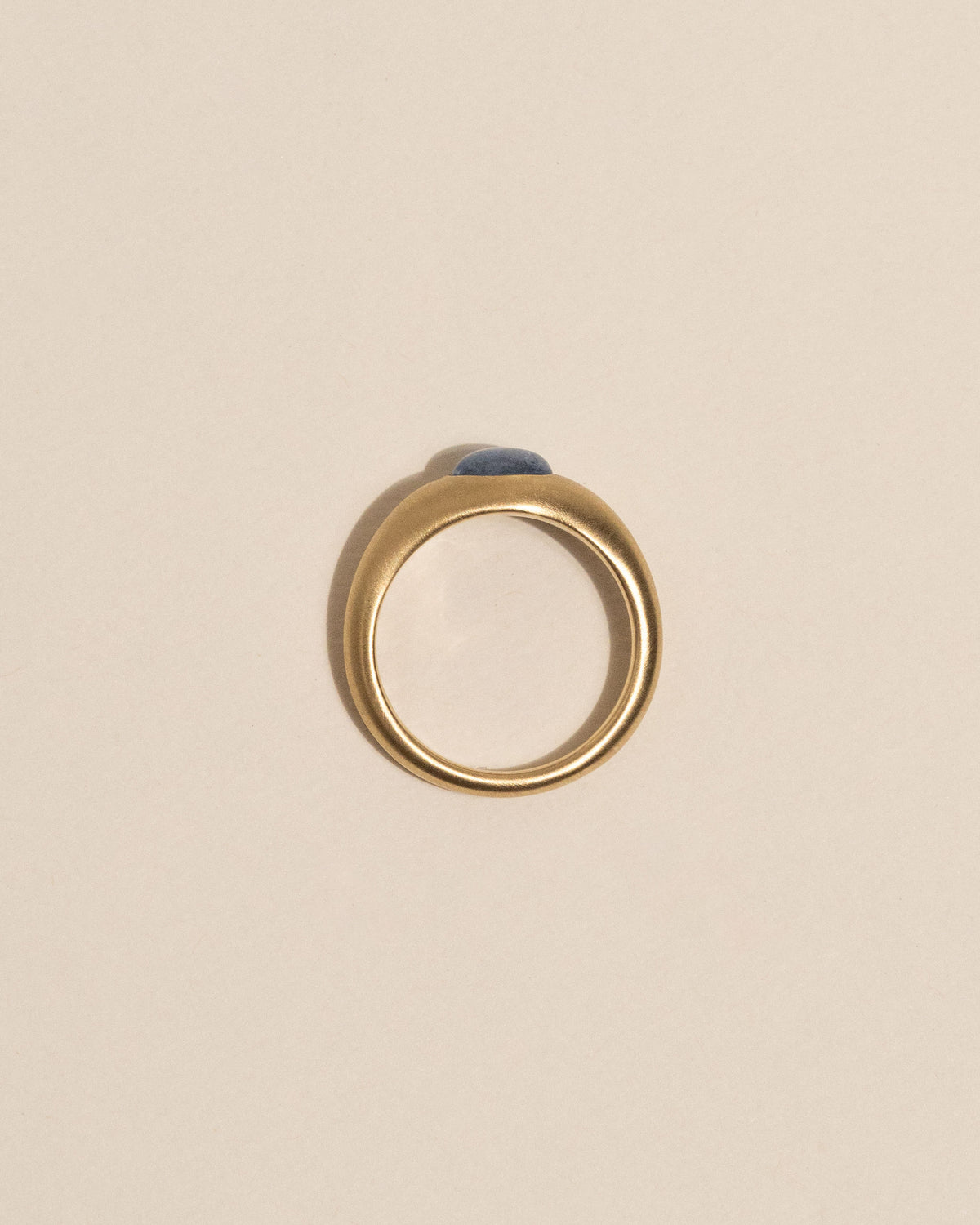 Top view of a gold signet ring with an oval blue stone, centered on a warm beige background.