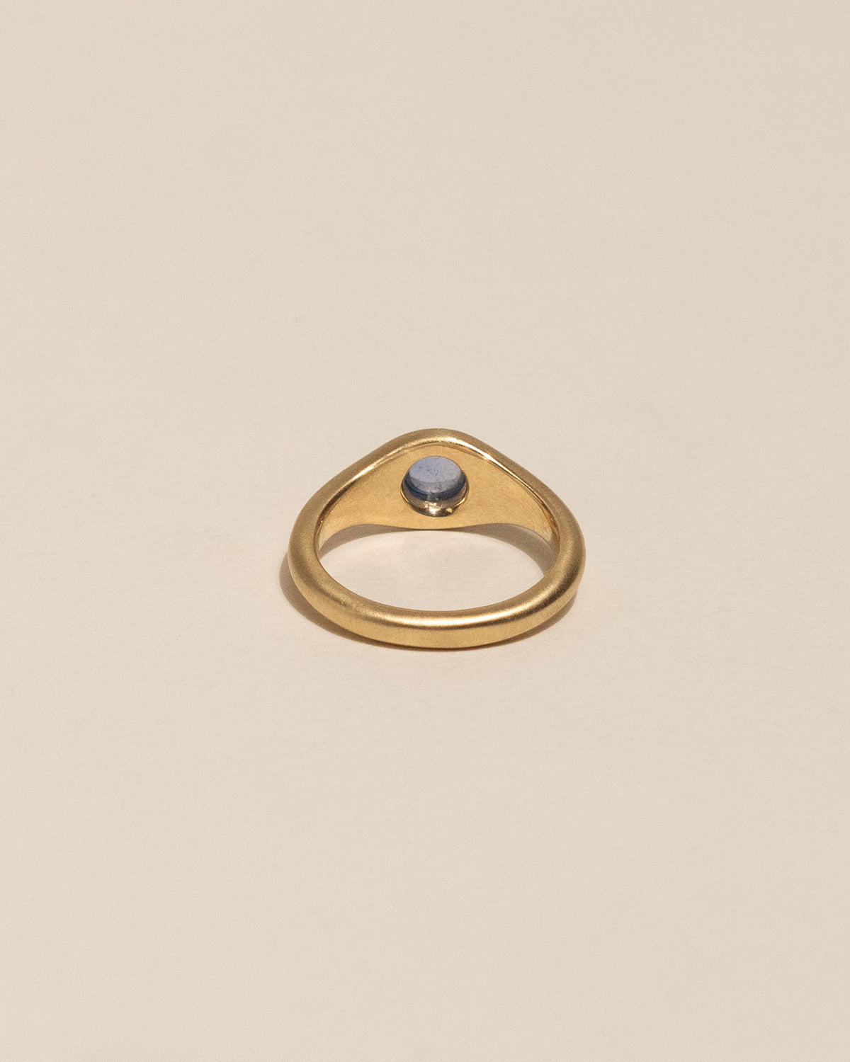 Back of a gold signet-style ring with a smooth, rounded bezel holding a pale blue oval gemstone, set against a solid warm beige background.