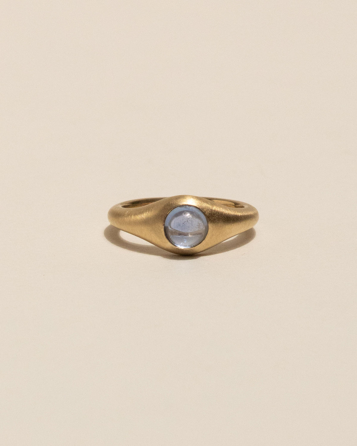 A gold signet-style ring with a smooth, rounded bezel holding a pale blue oval gemstone, set against a solid warm beige background.
