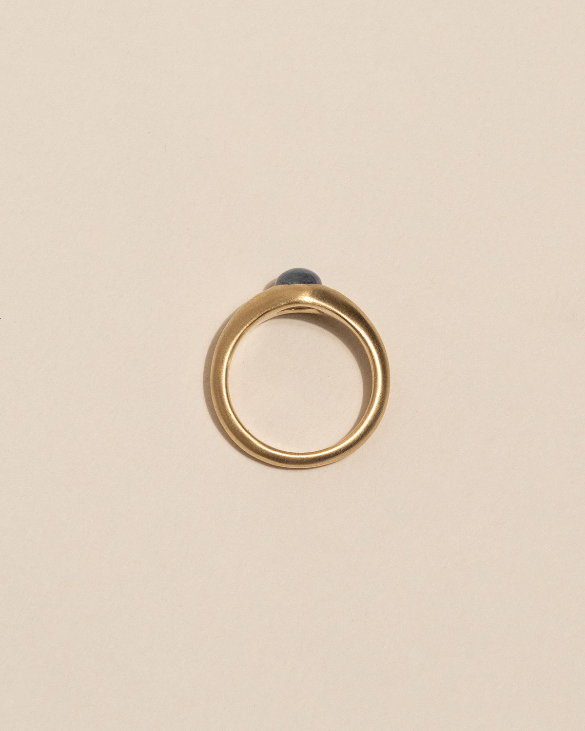 Top view of a gold signet-style ring with a smooth, rounded bezel holding a pale blue oval gemstone, set against a solid warm beige background.
