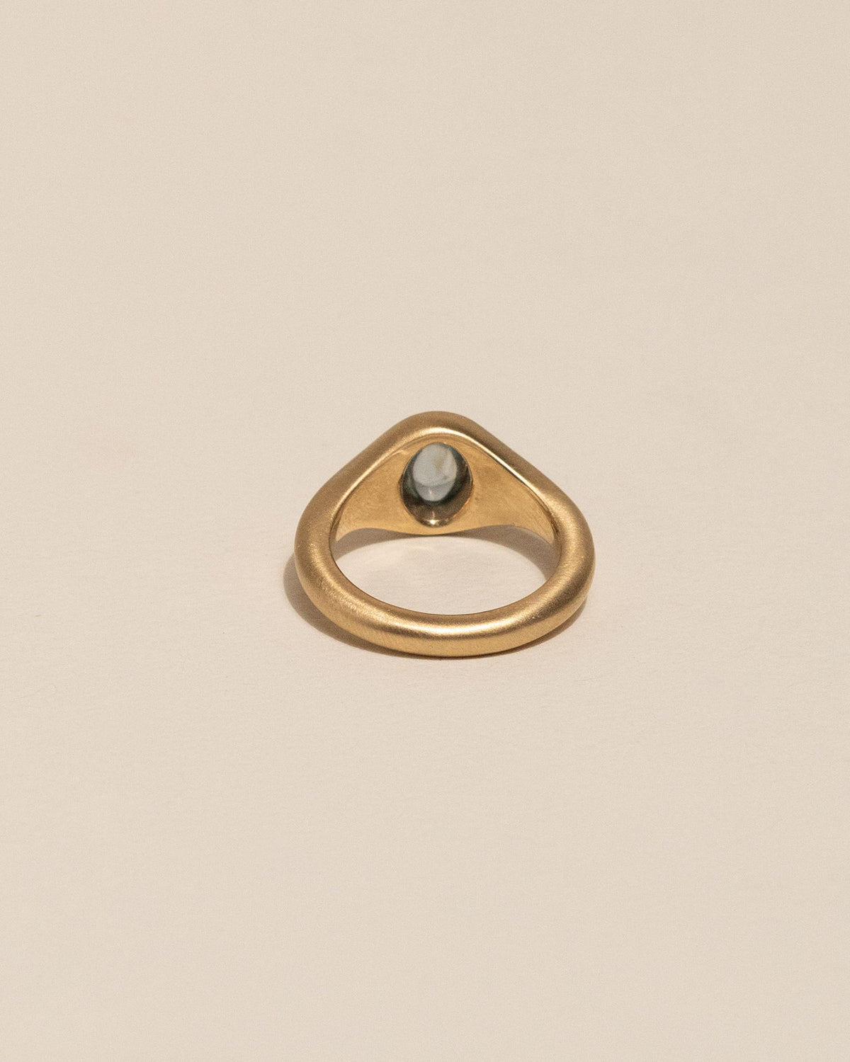 Back of a gold signet-style ring with a smooth, rounded blue sapphire in the center, displayed on a clean, solid beige background.
