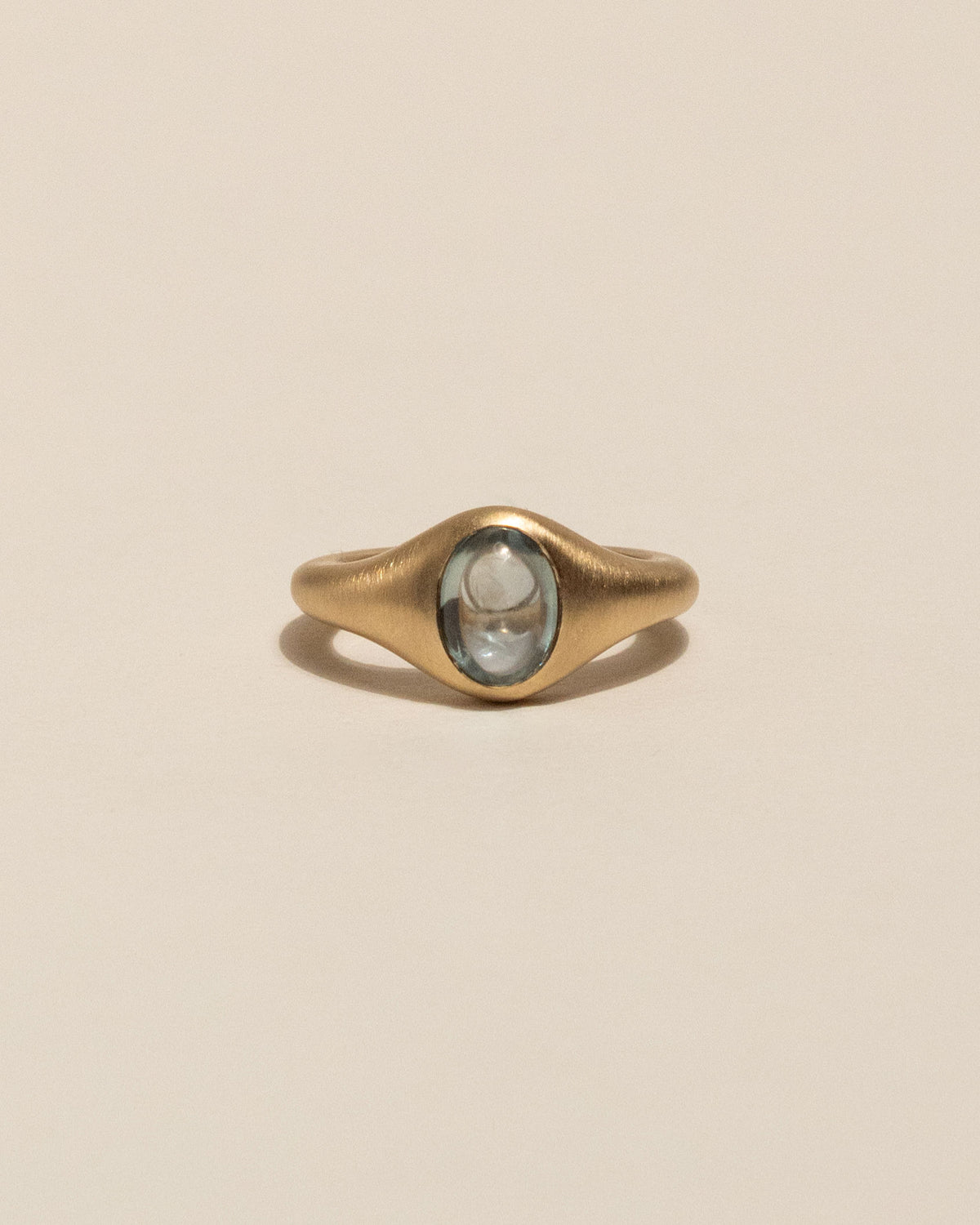 A gold signet-style ring with a smooth, rounded blue sapphire in the center, displayed on a clean, solid beige background.