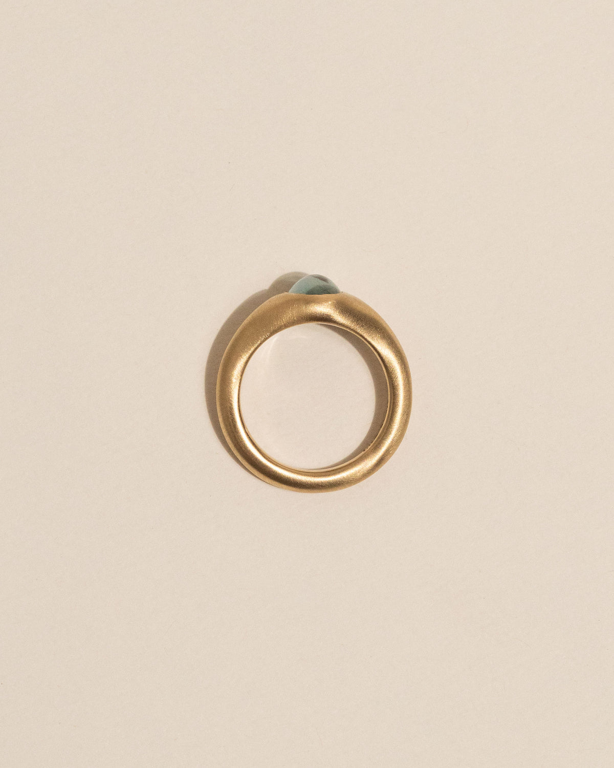 Top view of a gold signet-style ring with a smooth, rounded blue sapphire in the center, displayed on a clean, solid beige background.