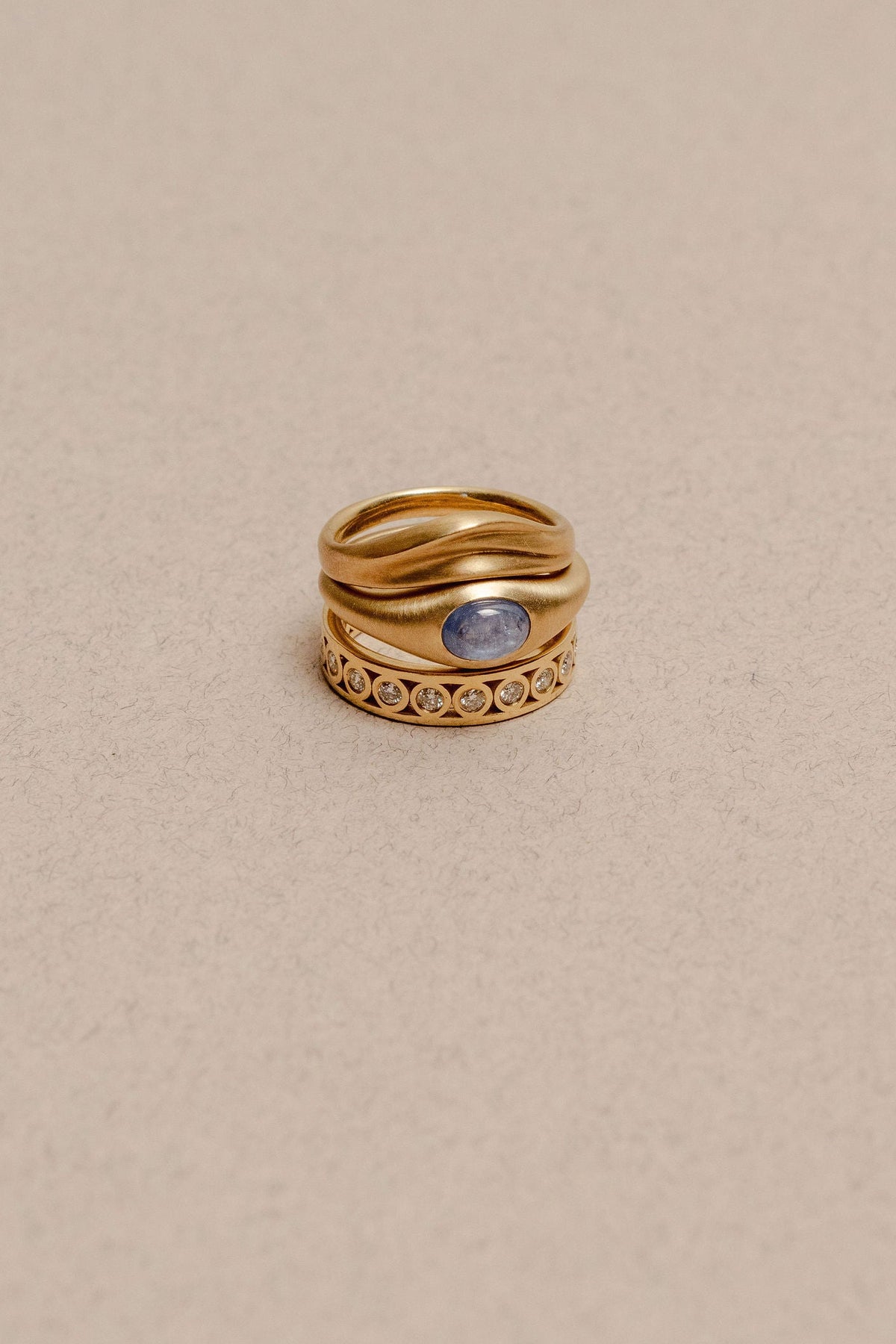 Three gold rings stacked together, one with a dune wavy texture band, a central oval blue sapphire, and a diamond eternity ring, photographed on a neutral beige background.