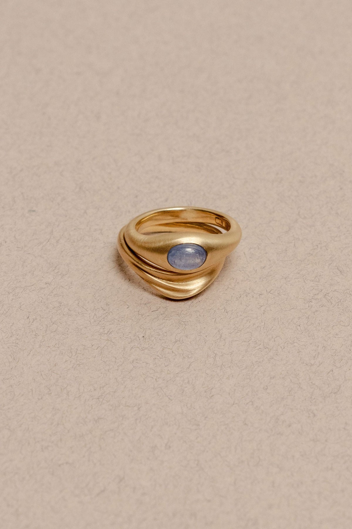 Two gold rings stacked together, one with a smooth surface and oval blue sapphire cabochon, the other is a curved gold ring with organic dune texture, displayed on a textured beige background.
