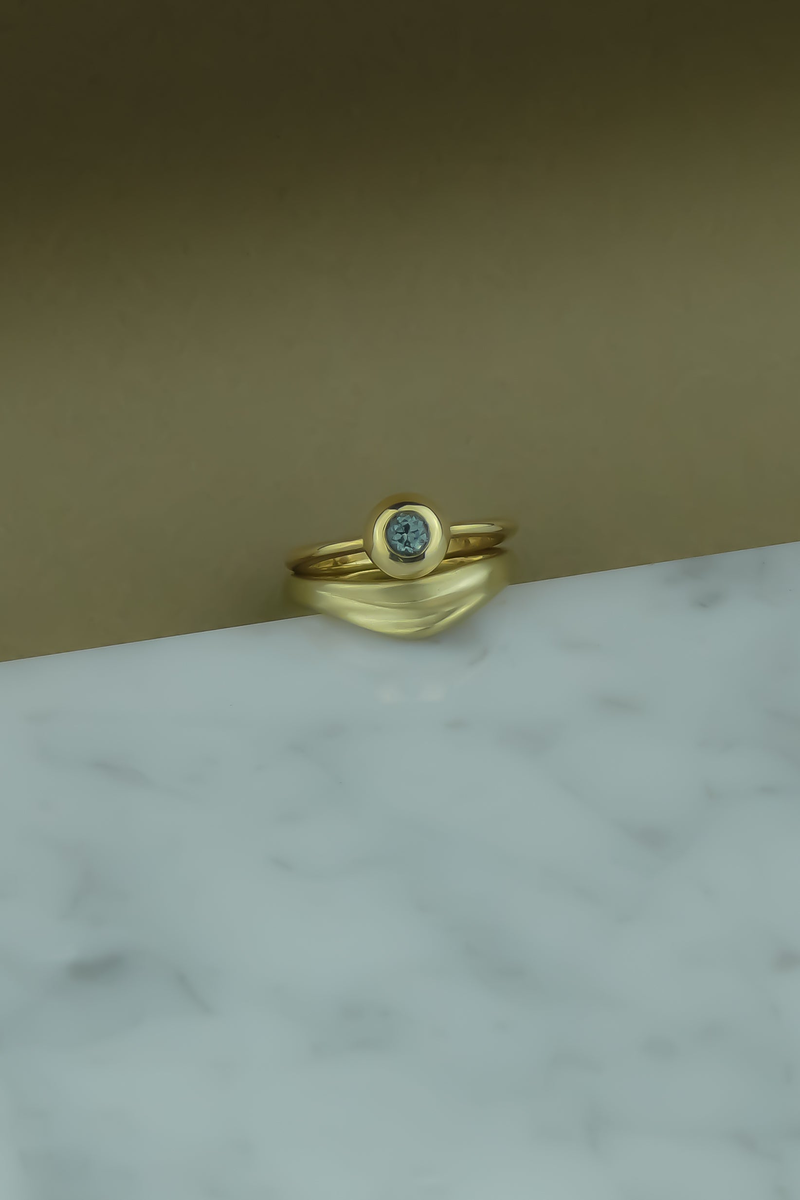 gold and teal sapphire foundation solitaire ring paired with a gold curved wedding band with organic texture on a marble background
