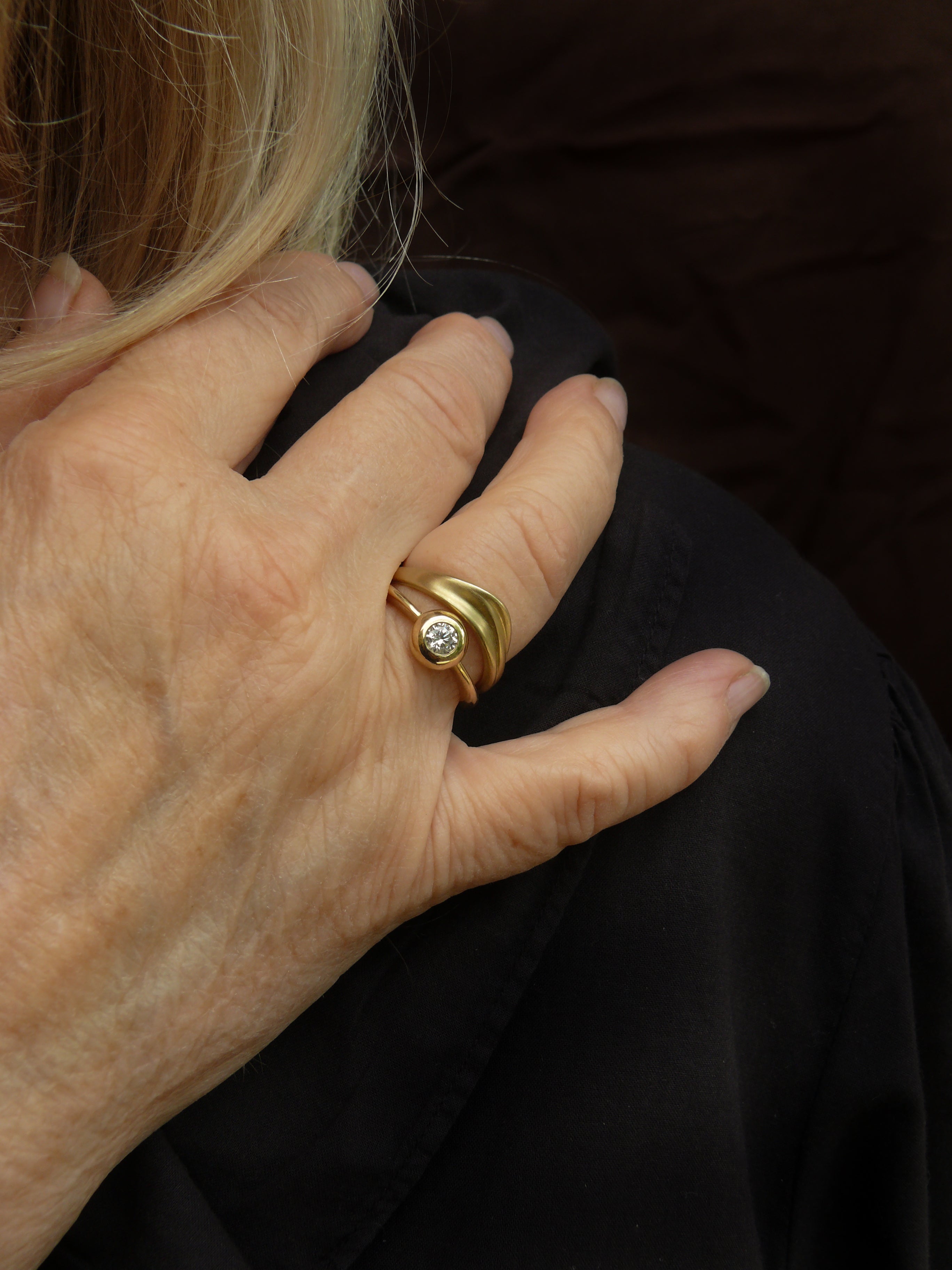 Close-up of a hand wearing a gold curved dune ring and a foundation solitaire ring with a diamond on a dark background