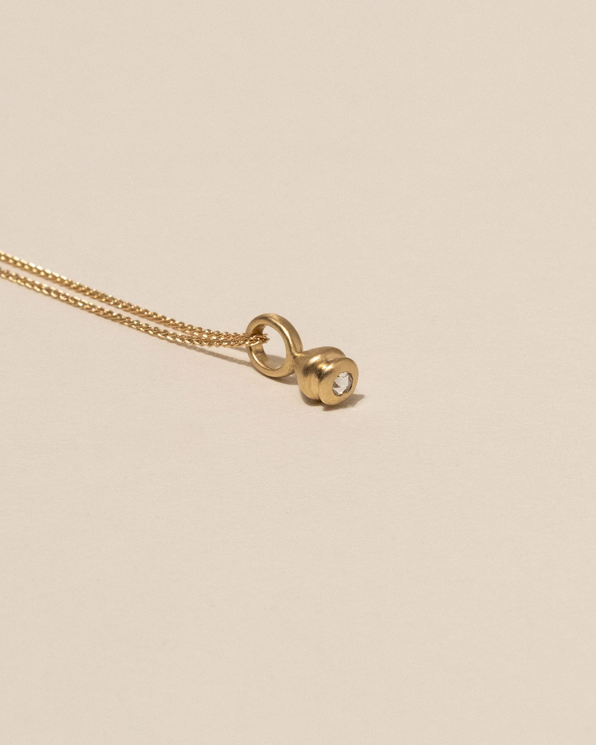 A gold pendant necklace featuring a teardrop-shaped pendant with a white Canadian rose cut diamond, displayed on a solid beige background.