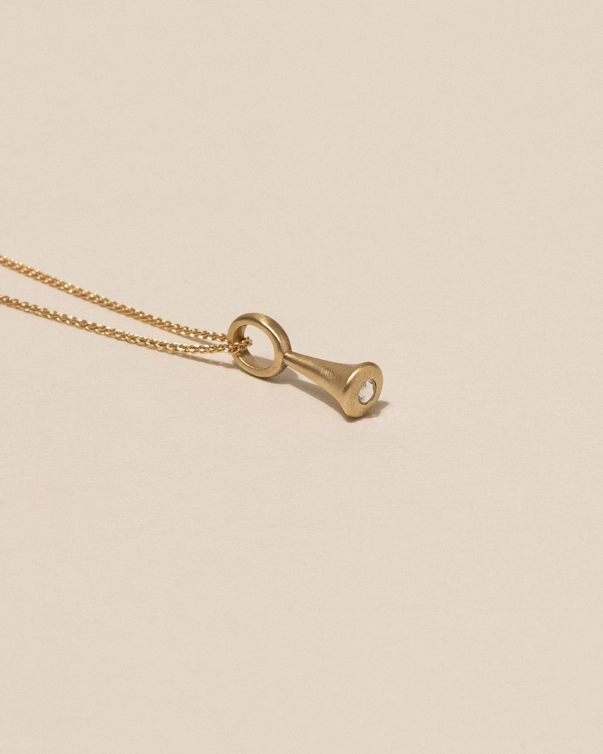 A gold pendant necklace featuring a teardrop-shaped pendant with a white Canadian rose cut diamond, displayed on a solid beige background.