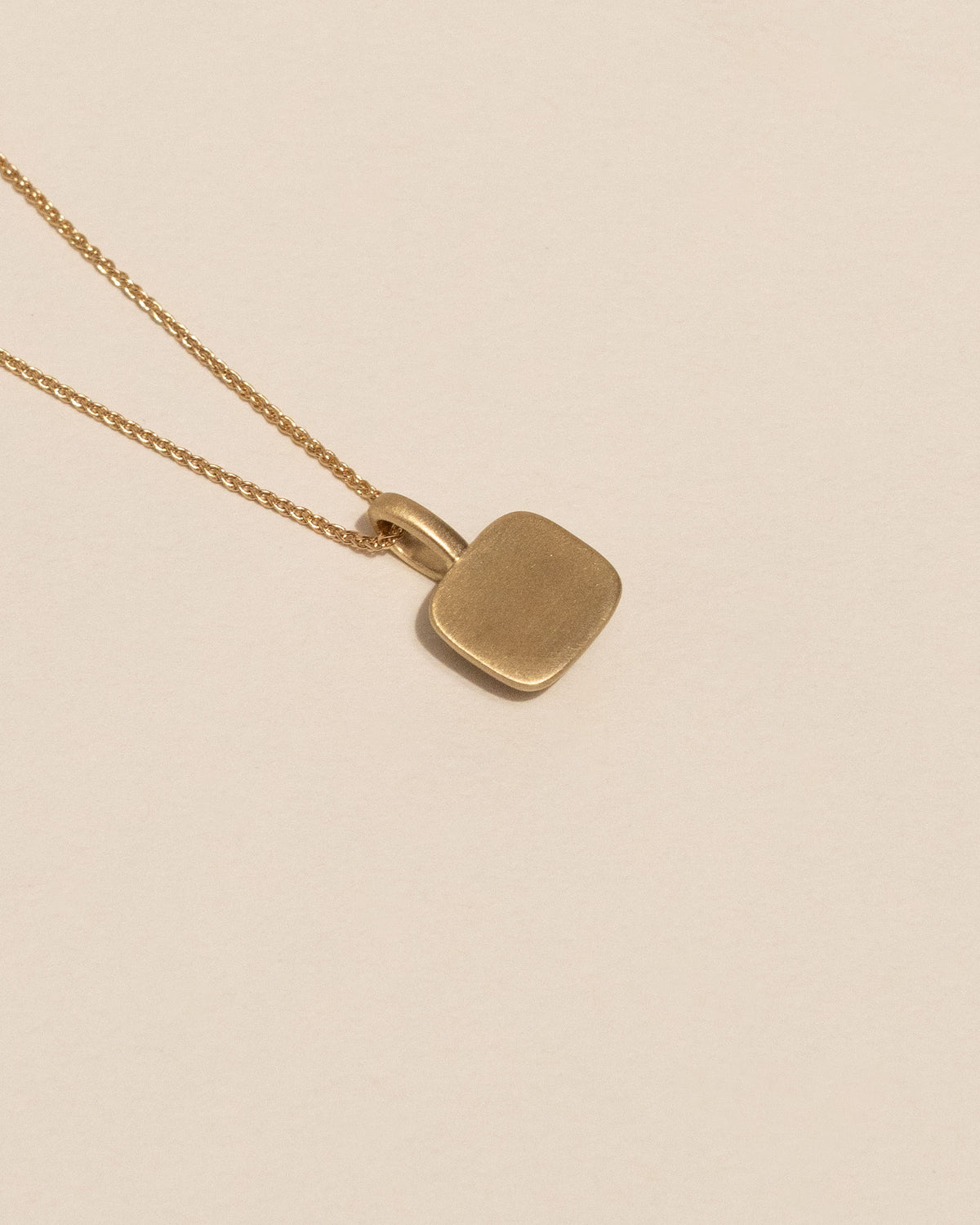 Back of a gold necklace with a small rounded square pendant featuring a single white diamond, displayed against a solid light beige background.