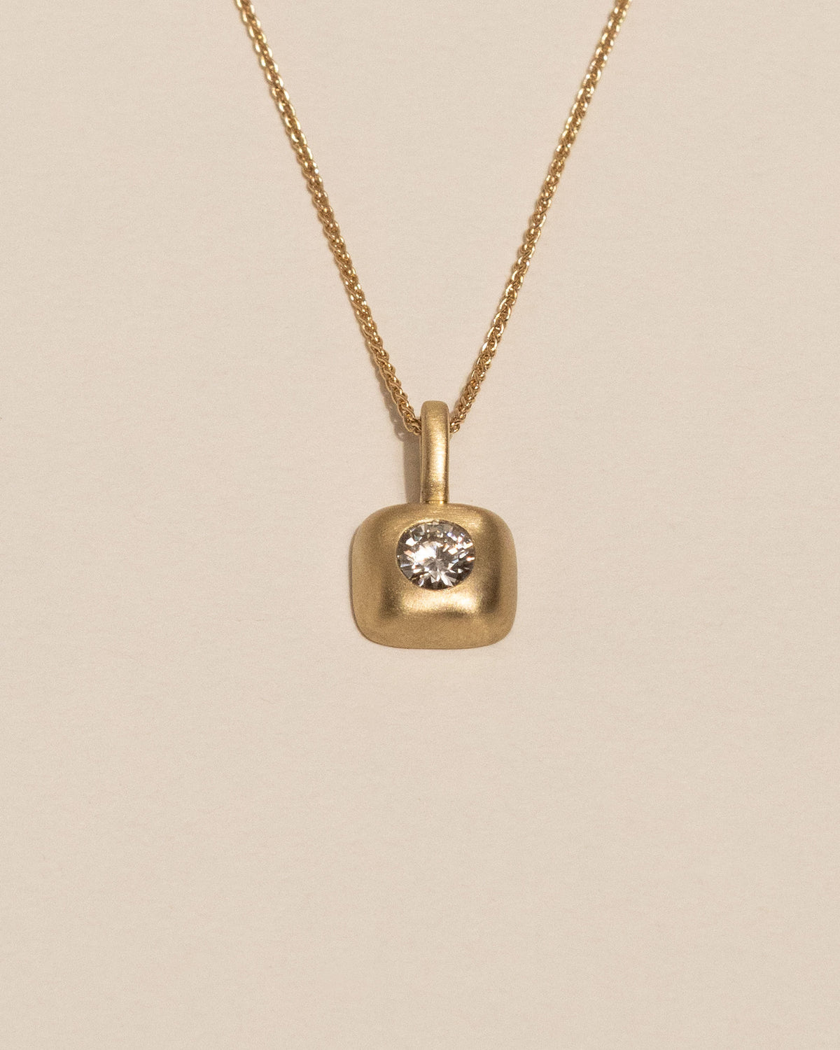 A gold necklace with a small rounded square pendant featuring a single white diamond, displayed against a solid light beige background.