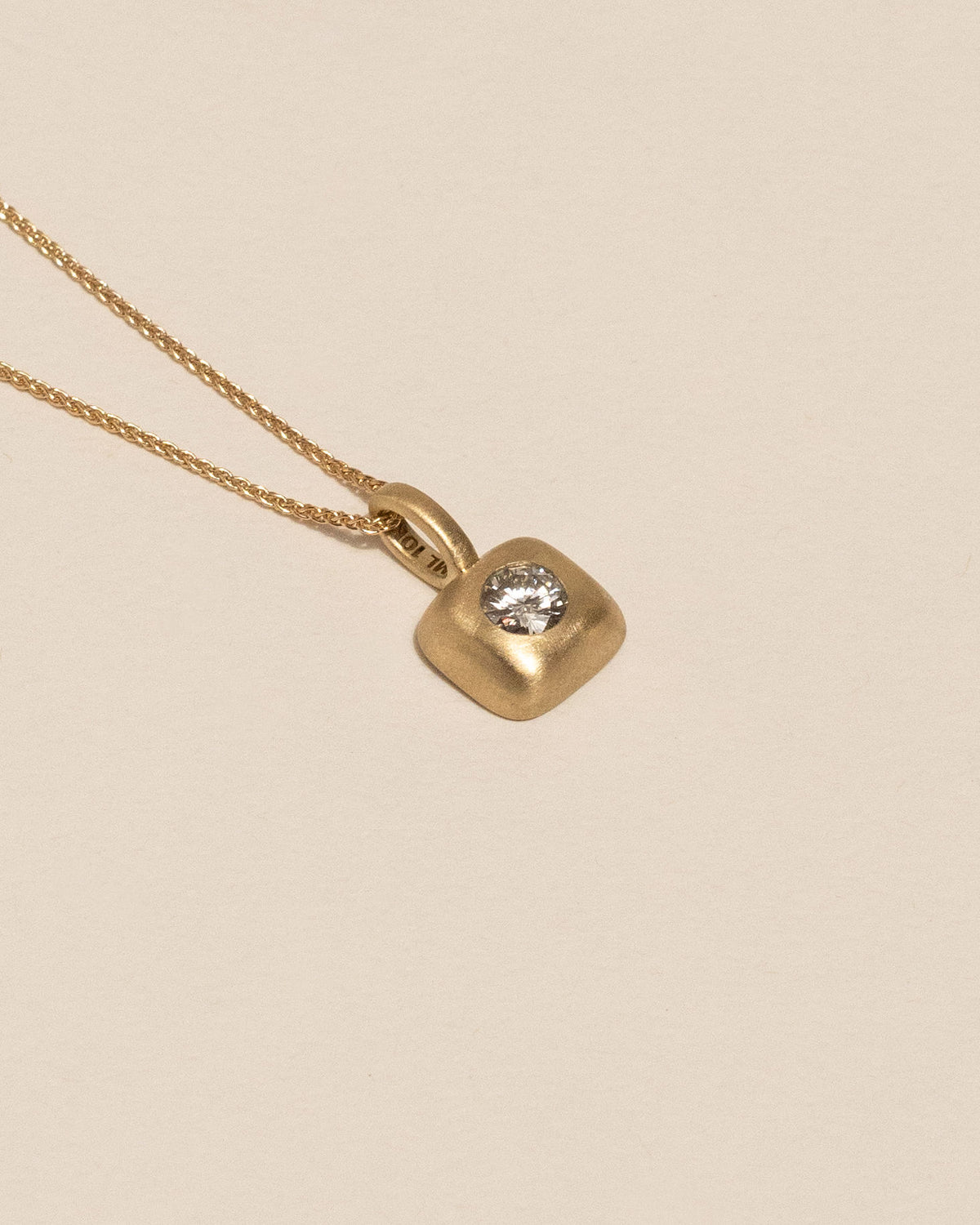 Side view of a gold necklace with a small rounded square pendant featuring a single white diamond, displayed against a solid light beige background.