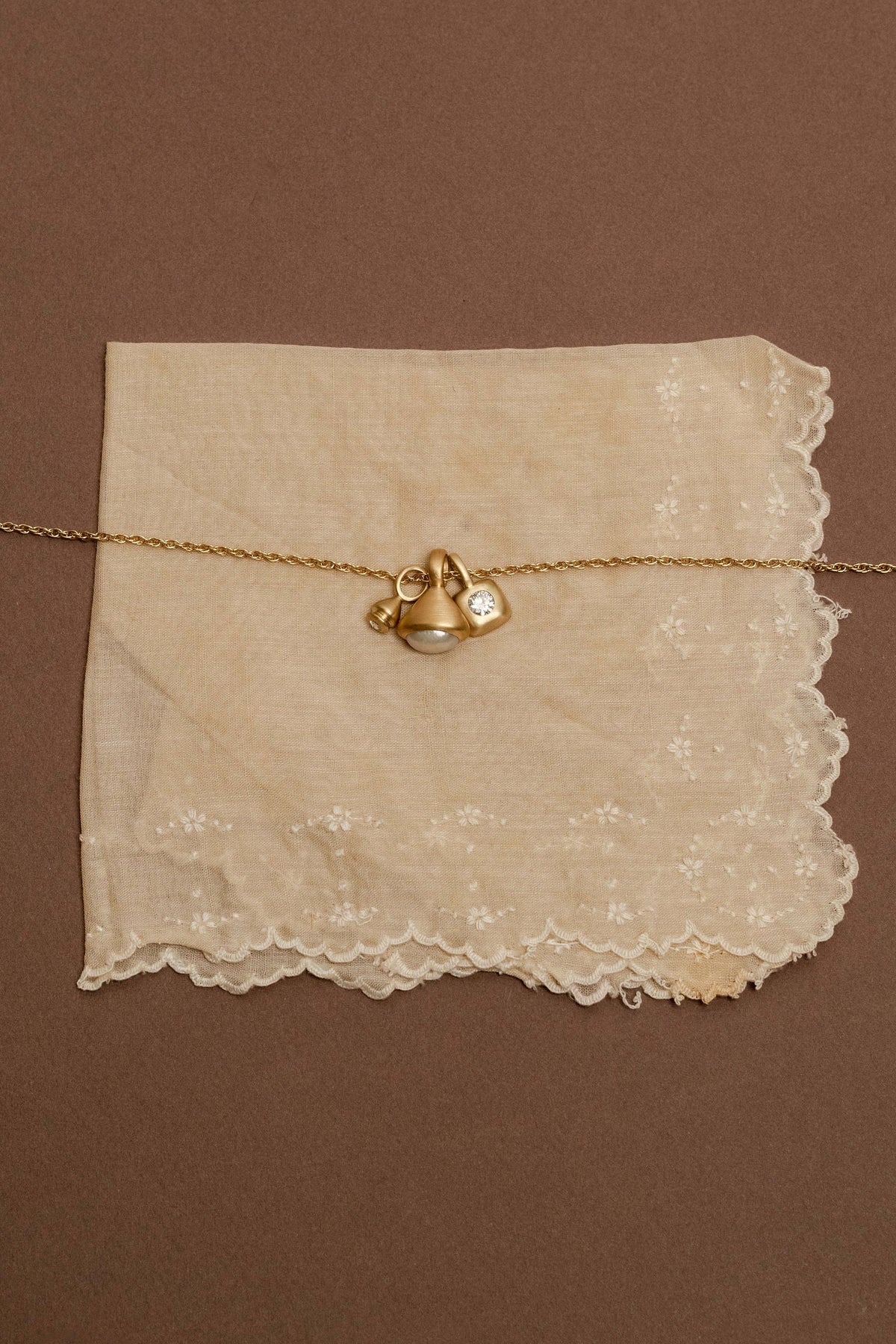 A gold rope chain with charms with diamonds and pearls, displayed on a square cream lace napkin against a solid brown background.