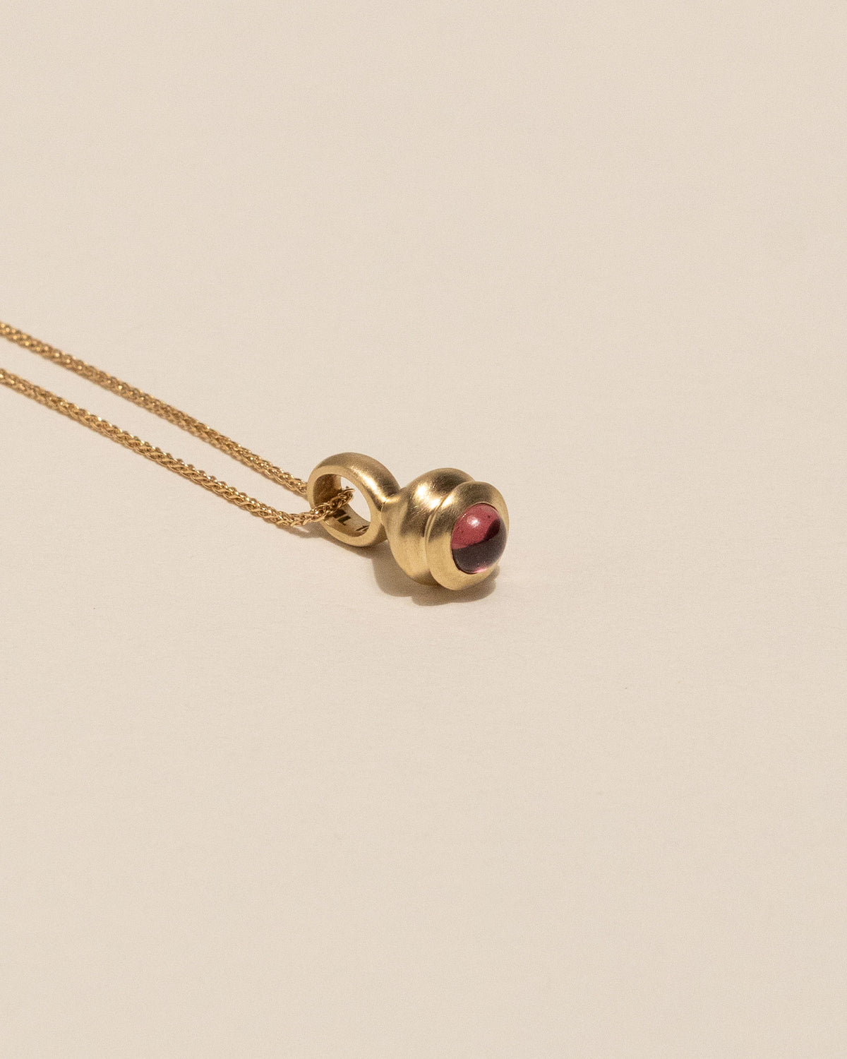 A gold chain necklace with a unique spherical pendant featuring a gold body and a small red garnet, displayed on a clean beige background.