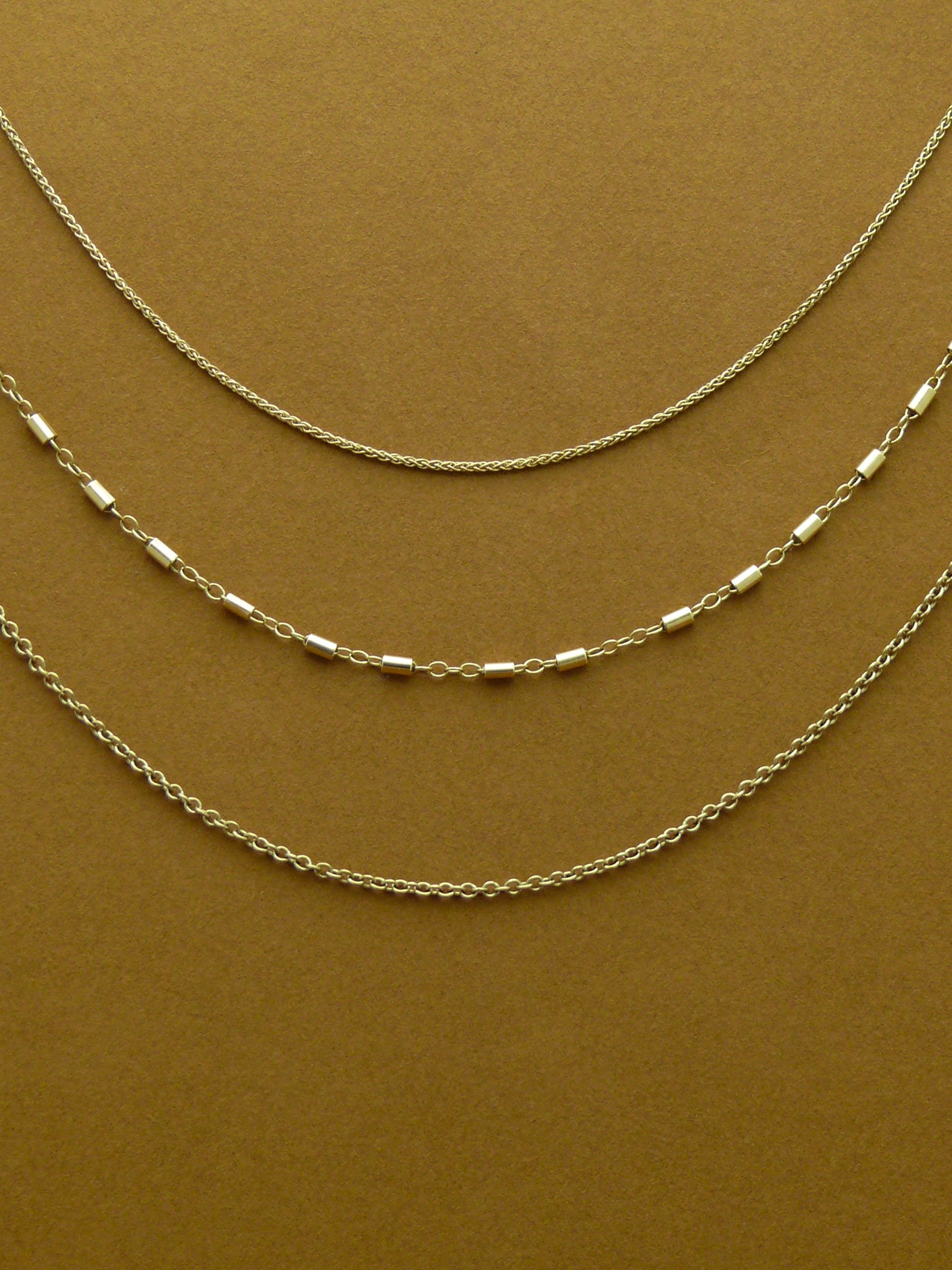 Three gold chains of different styles on a brown background, including the heritage wheat chain, stellar bar chain, and strand cable chain