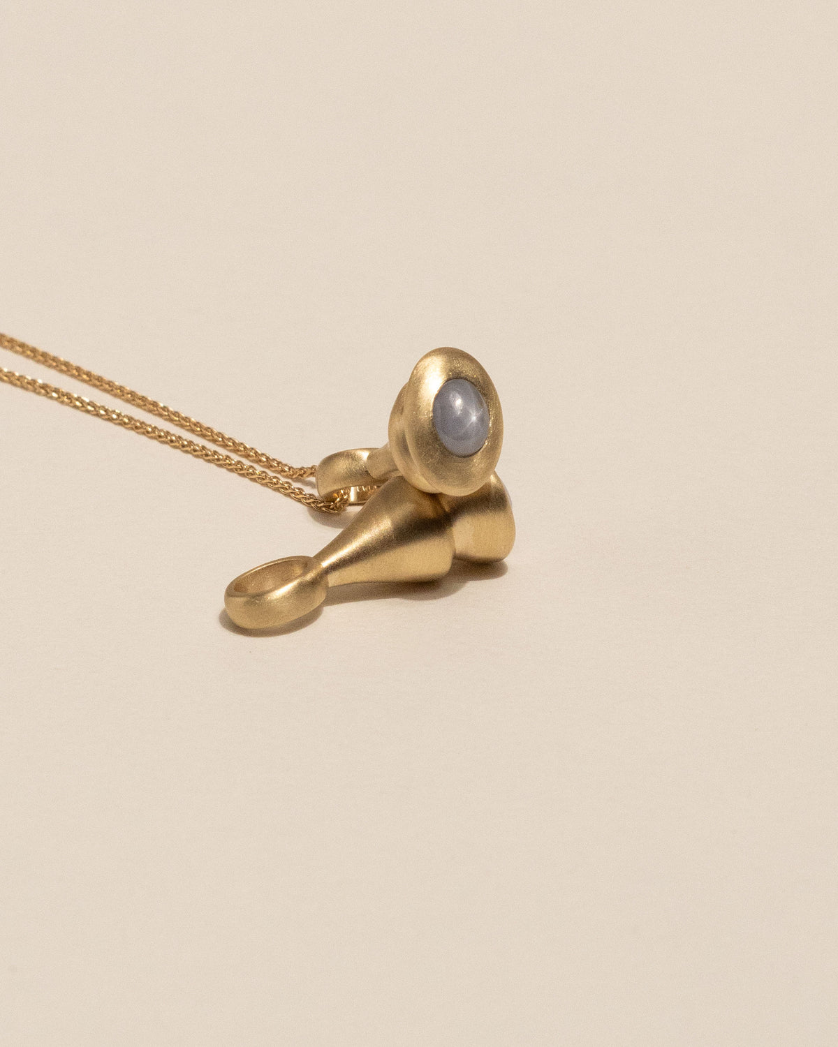 A gold pendant necklace featuring a small, bulbous gold charm with a smooth, oval light lavendar-gray star sapphire bezel set, resting on a warm beige background.