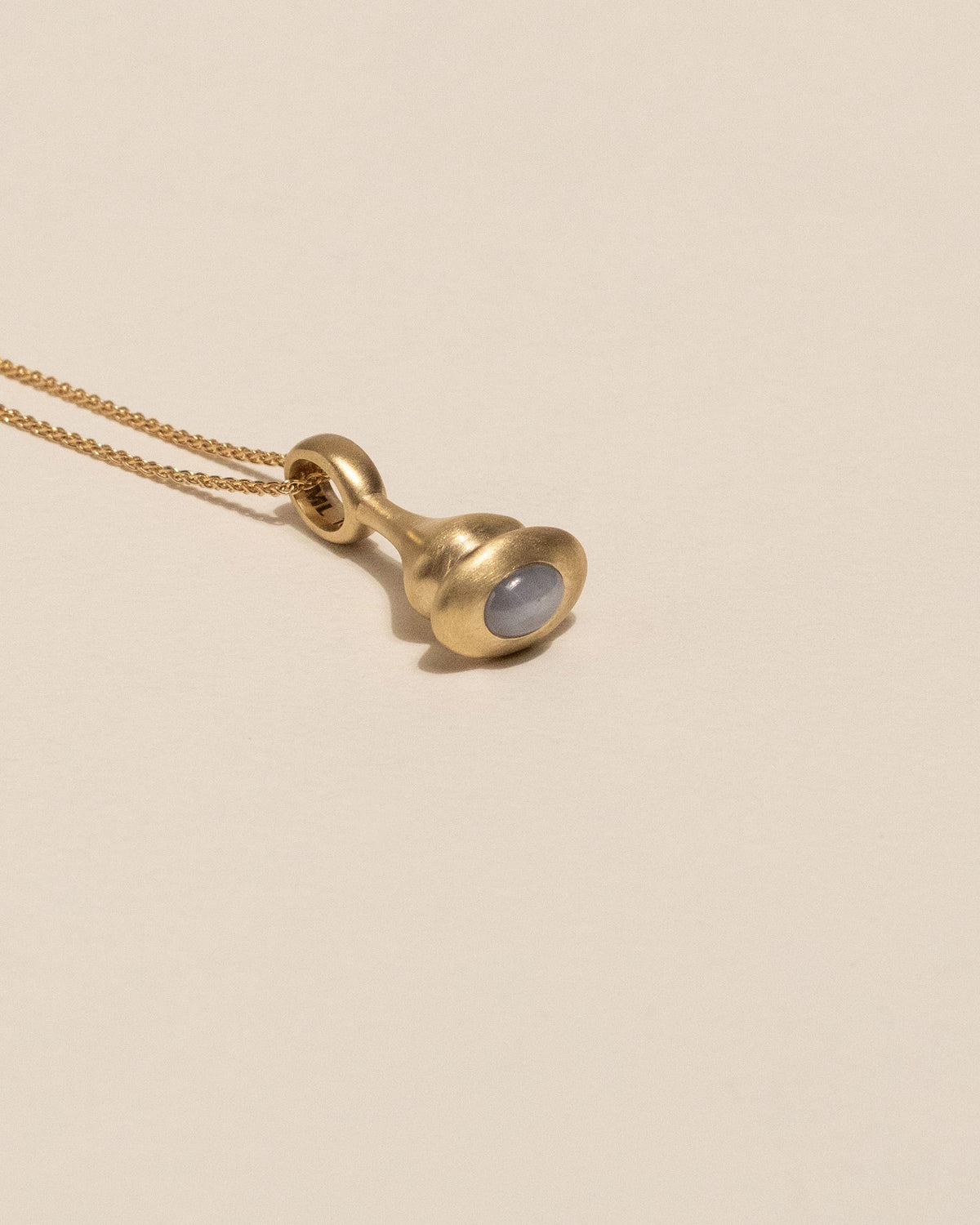 A gold pendant necklace featuring a small, bulbous gold charm with a smooth, oval light lavendar-gray star sapphire bezel set, resting on a warm beige background.