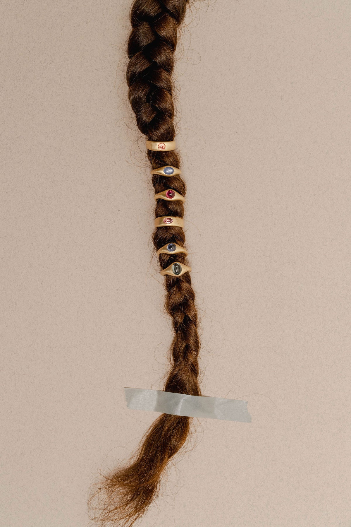 A close-up of a long brown braided hair, secured with tape near the bottom, decorated with several gold sapphire rings featuring colorful gemstones, against a plain beige background.