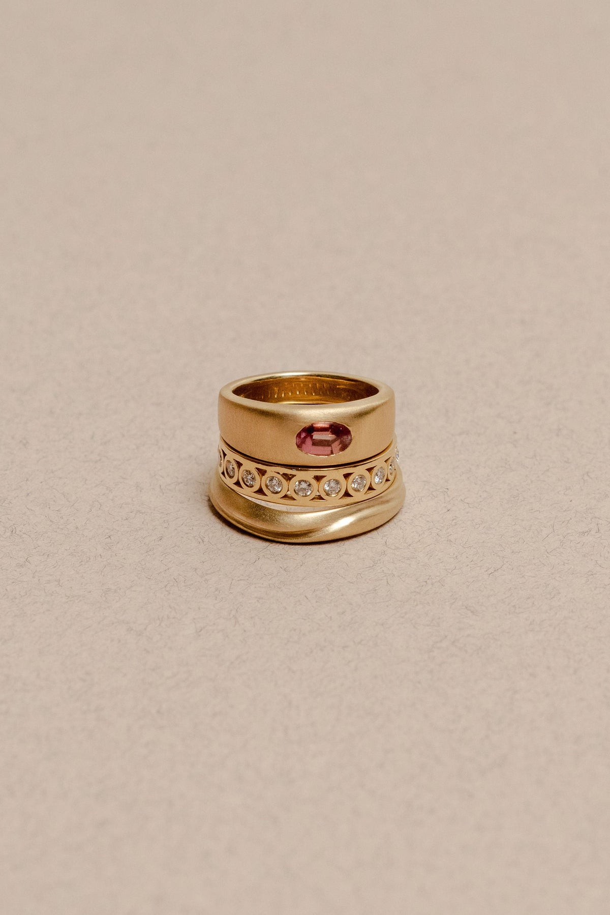 A gold ring with a central oval pink gemstone and a row of small clear stones, photographed on a textured beige background.