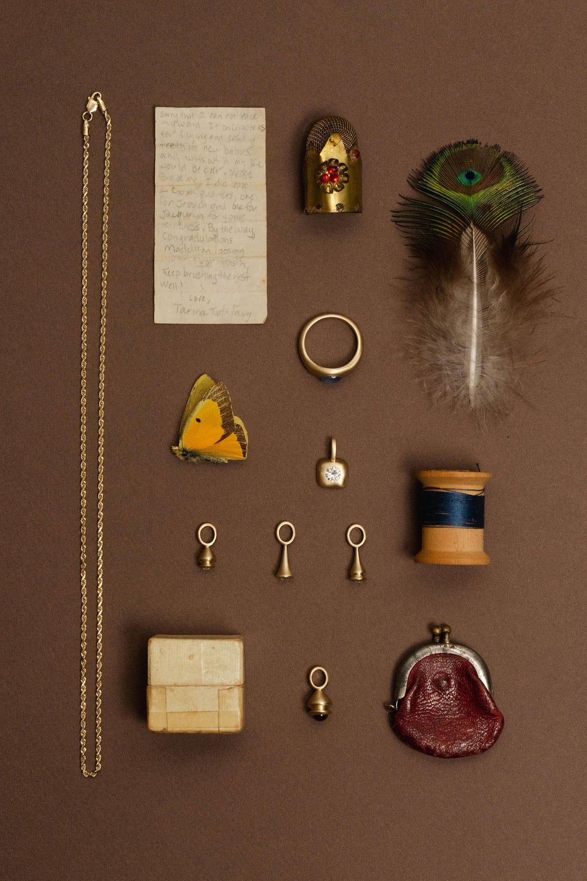 A curated flat-lay of jewelry and vintage objects arranged on a solid brown background, including a gold chain necklace, a peacock feather, a gold ring, pendulum charms, a small box, and other antique pieces.