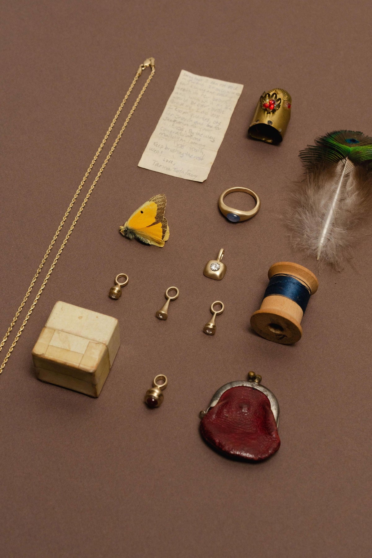 A curated flat-lay of jewelry and small accessories on a brown background, including a gold chain, rings, a butterfly, pendulum charms, a feather, thread spool, coin purse, and small boxes.