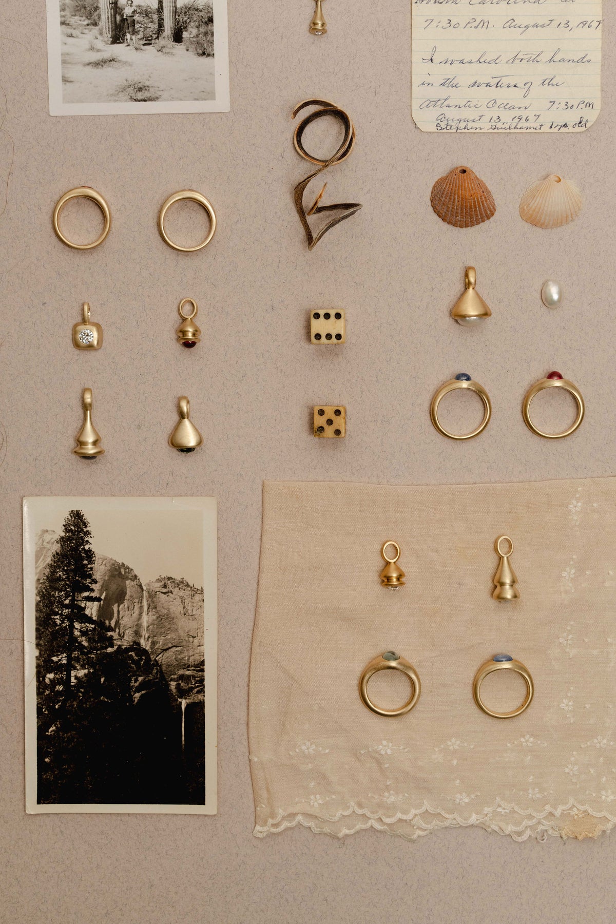 A flat-lay of jewelry and objects featuring multiple gold rings, small jewelry charms, shells, a pearl, and vintage photographs arranged on a beige paper.