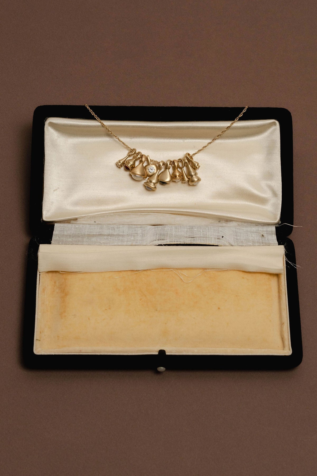 A gold necklace with a cluster of small charms, displayed inside an open vintage black jewelry box with cream satin lining, set against a solid brown background.