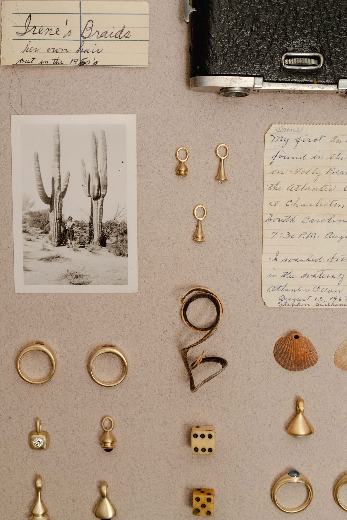 A styled flat-lay of gold jewelry findings and small accessories arranged on a beige fabric background, including several gold and sapphire rings, earrings, pendulum charms, a shell, dice, handwritten notes, and a black-and-white cactus photo.