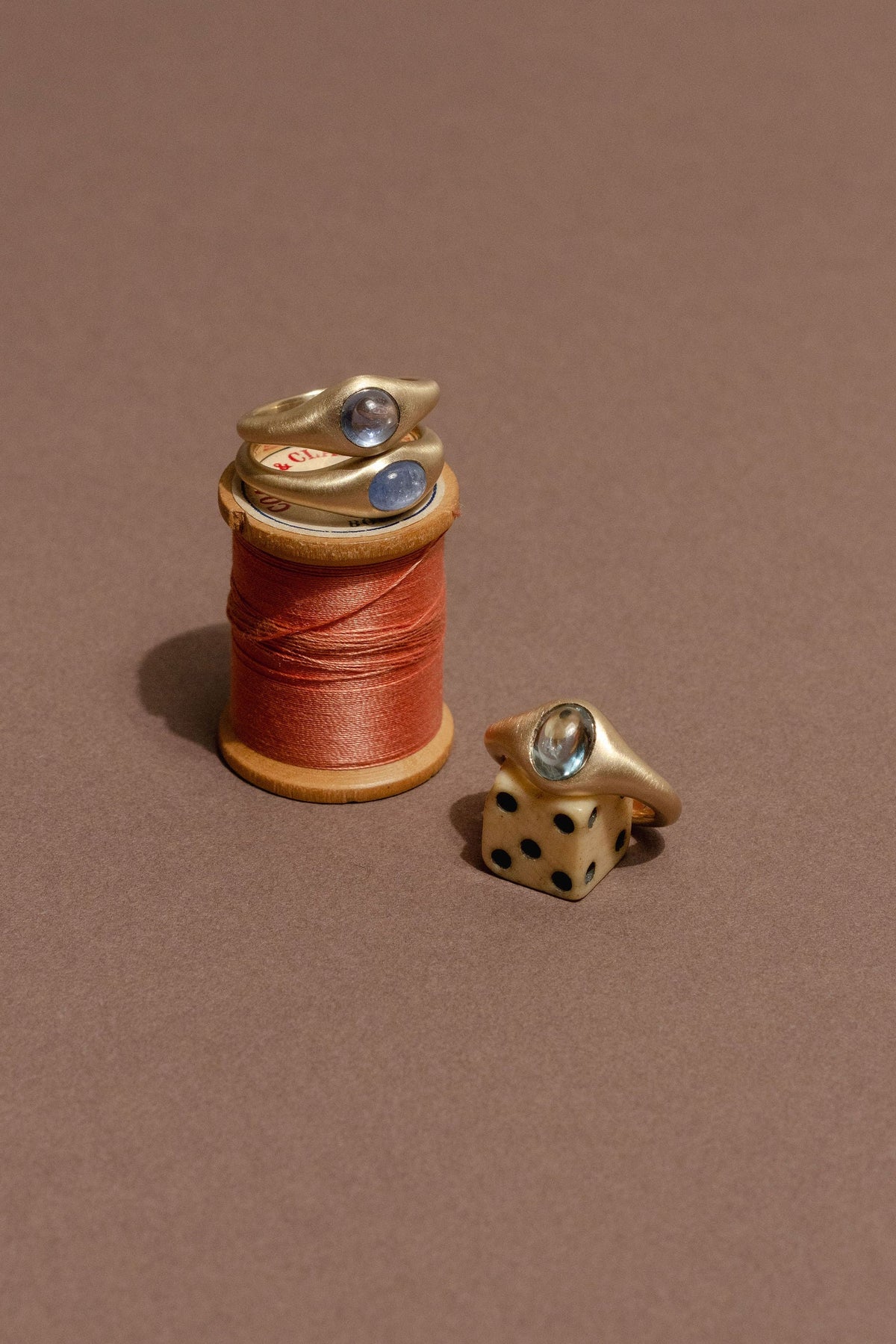 Three gold rings with blue sapphires, two of the resting on a spool of pink thread and the other resting on a vintage dice, on a neutral brown backround.