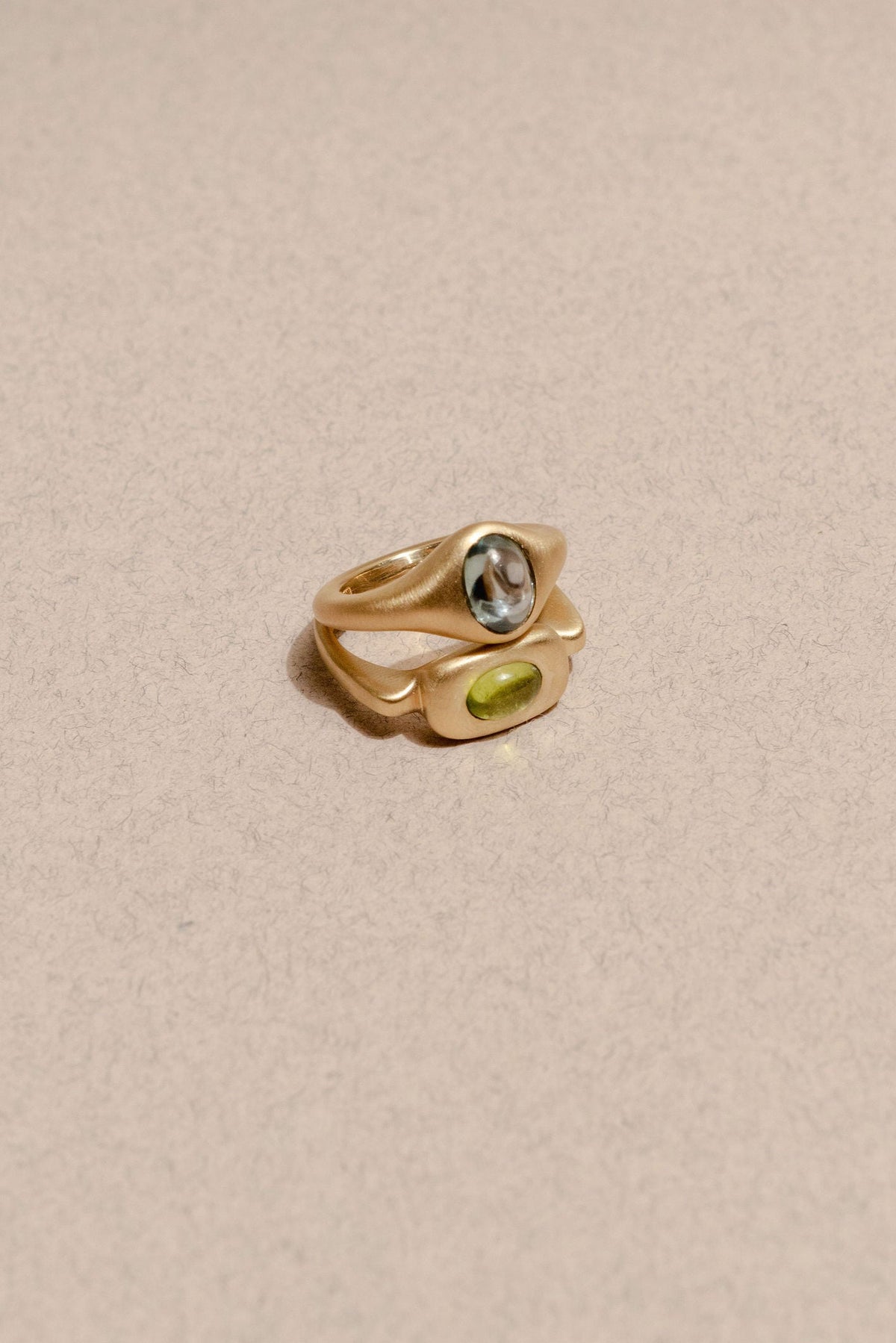 Two gold rings stacked, one with a blue sapphire set north to south and one with a green peridot set east to west, resting on a neutral background. 