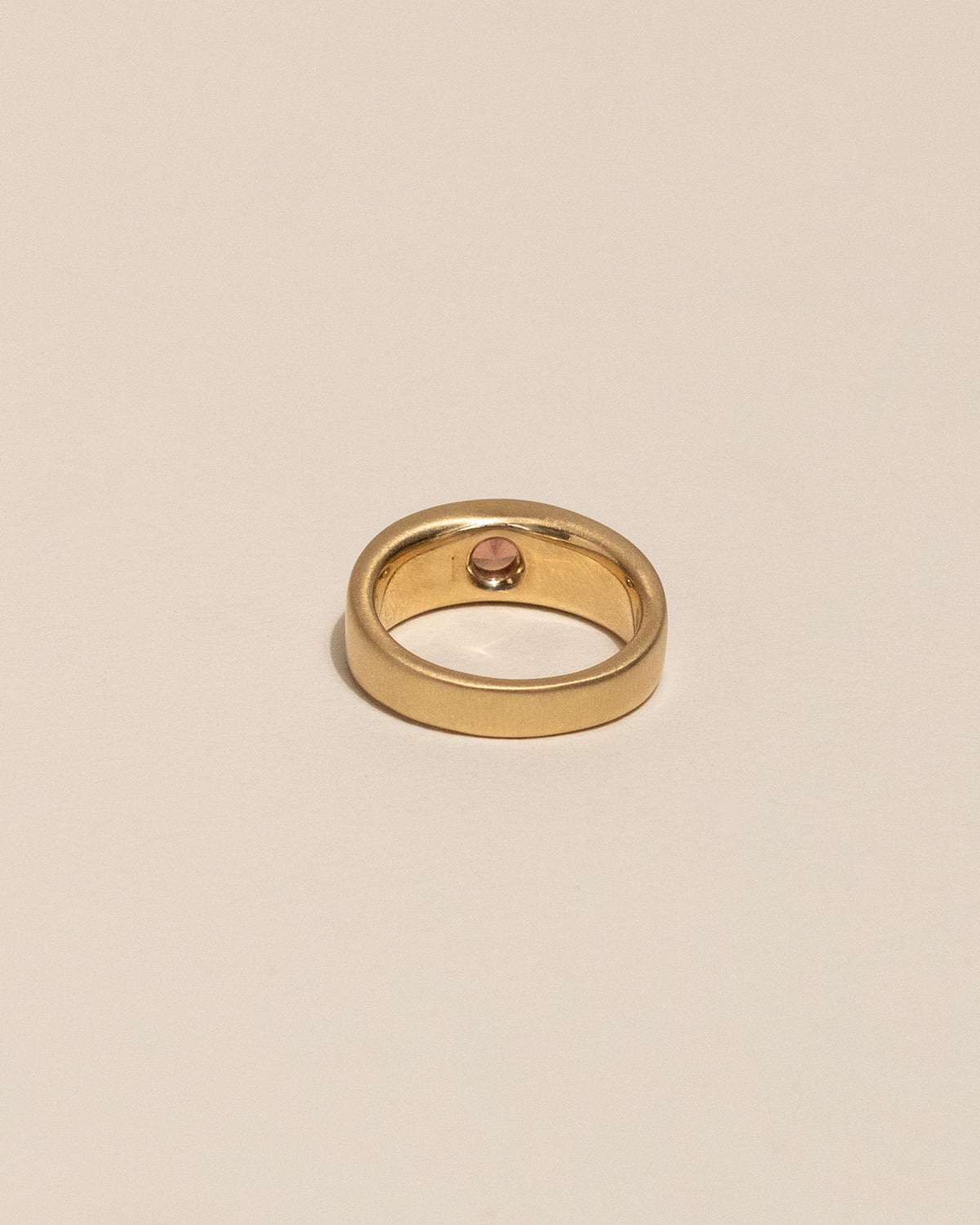 Back of a gold plateau ring with a small peach buff top sapphire flush set into a flat gold band, centered on a plain beige background.