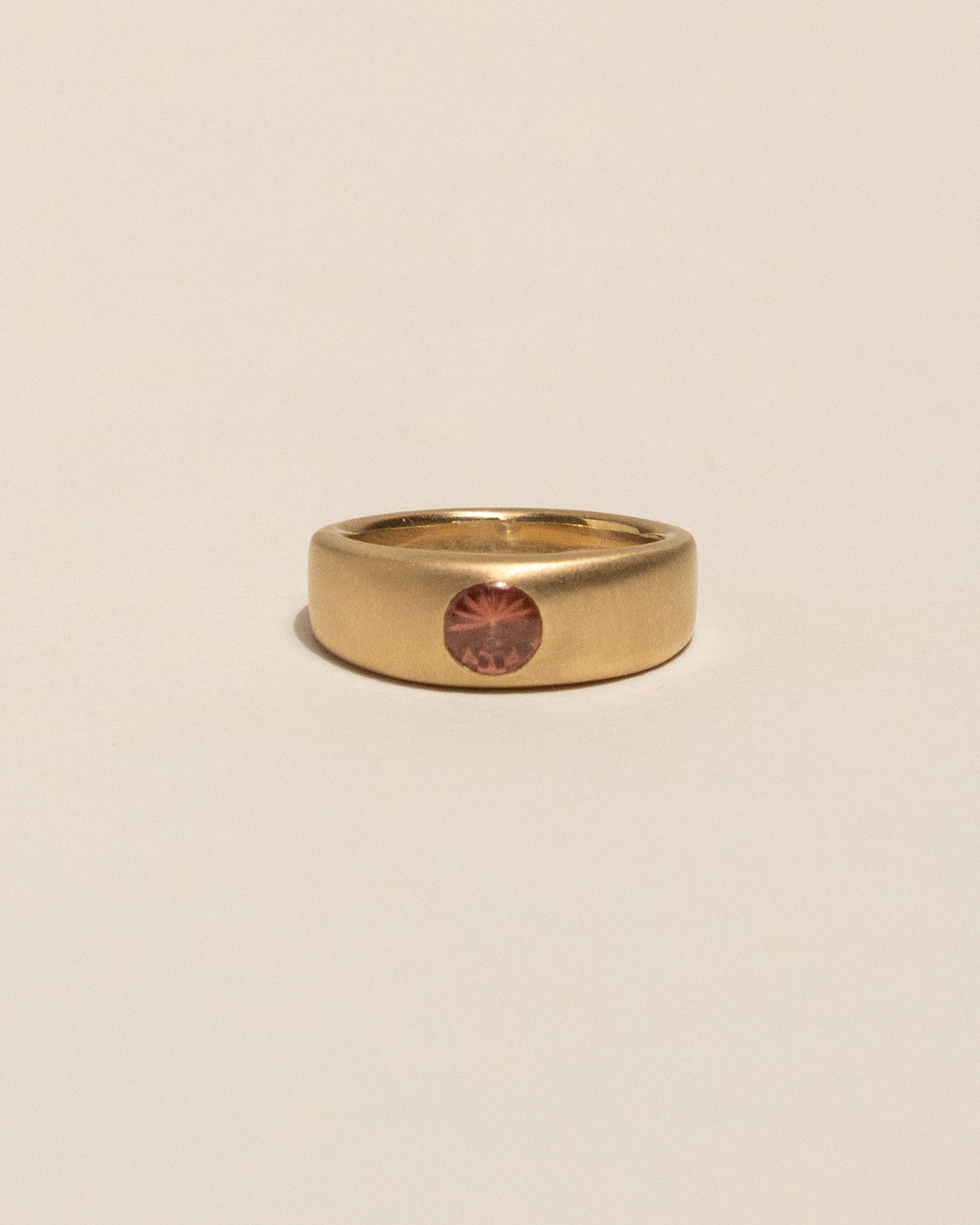 A gold plateau ring with a small peach buff top sapphire flush set into a flat gold band, centered on a plain beige background.