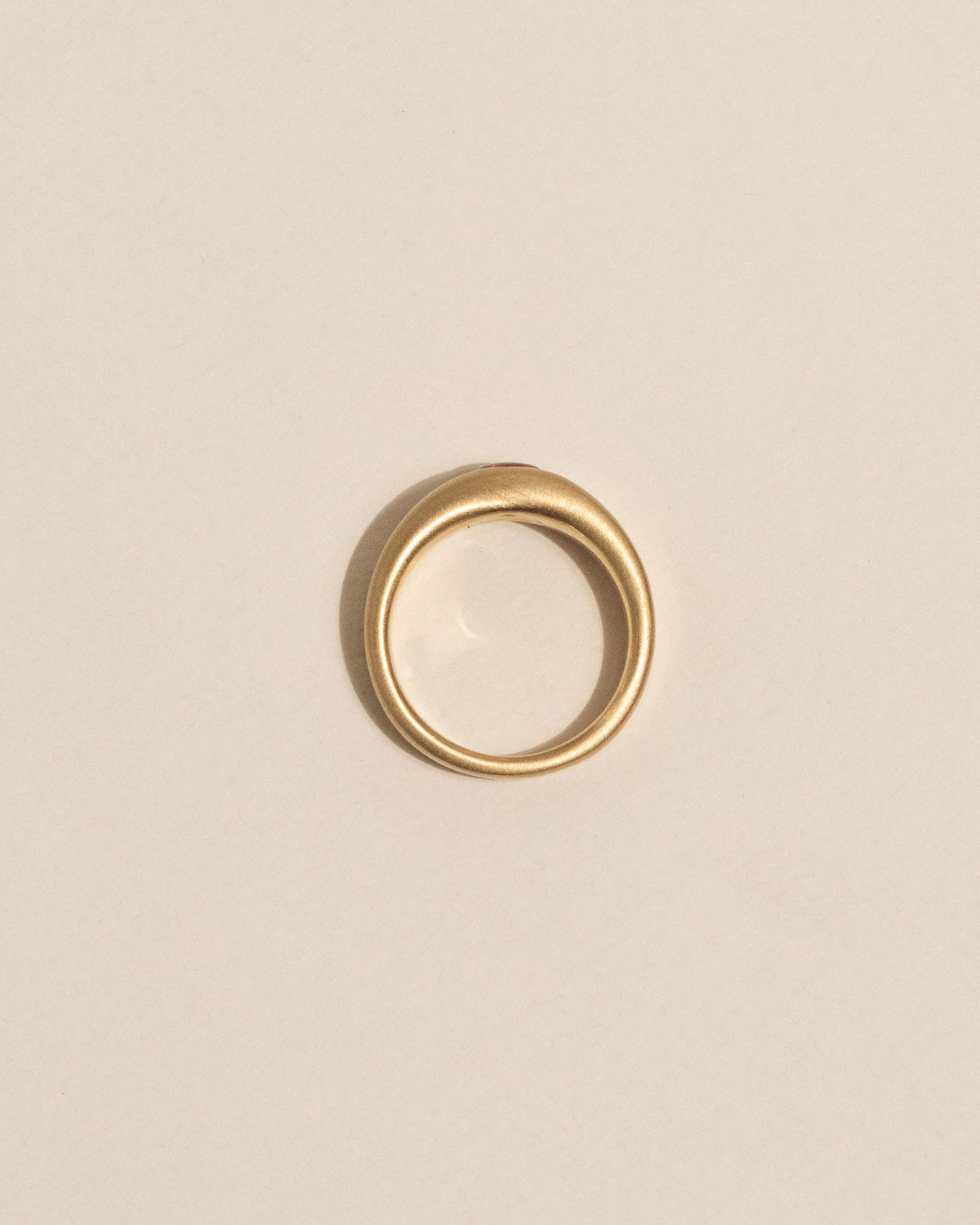 Top view of a gold plateau ring with a small peach buff top sapphire flush set into a flat gold band, centered on a plain beige background.