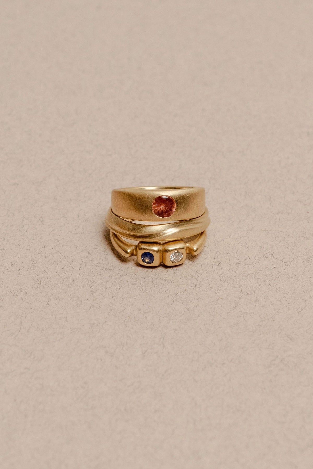 A gold plateau ring with a small peach buff top sapphire flush set into a flat gold band, centered on a plain beige background, stacked with 2 other gold rings with dune texture and flush set gemstones.